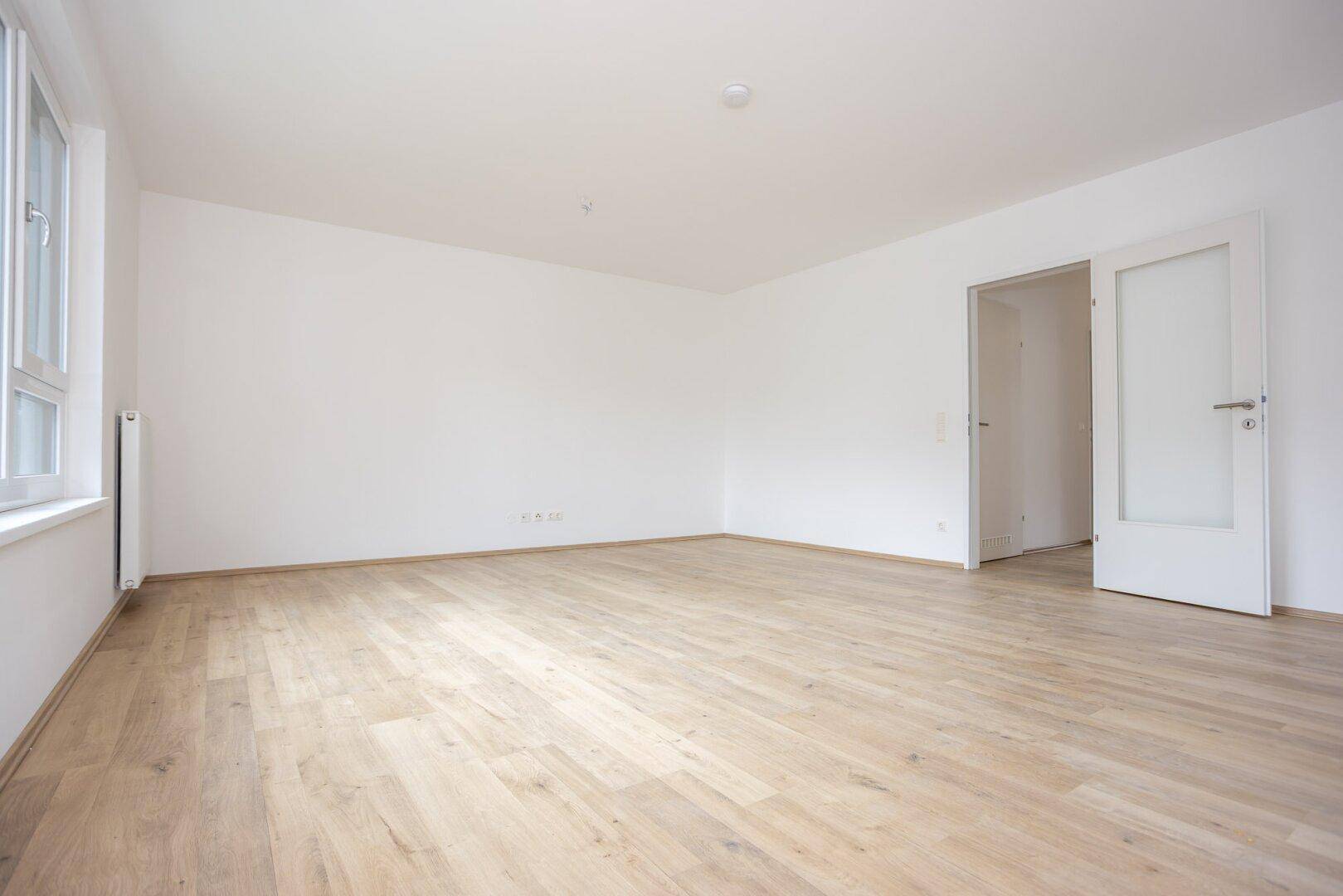 Großzügiger Wohnraum mit hellem Laminatboden, weißen Wänden und einem großen Fenster.