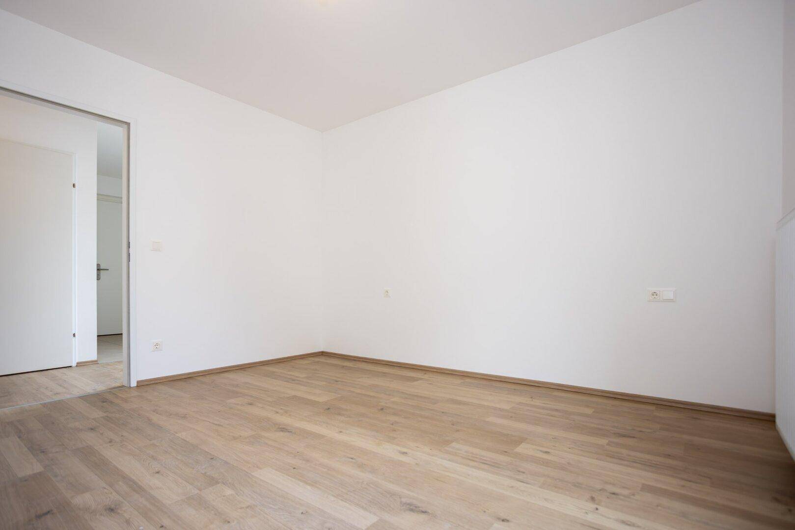 Leeres Zimmer mit hellem Laminatboden und weißen Wänden, bereit zur individuellen Gestaltung.