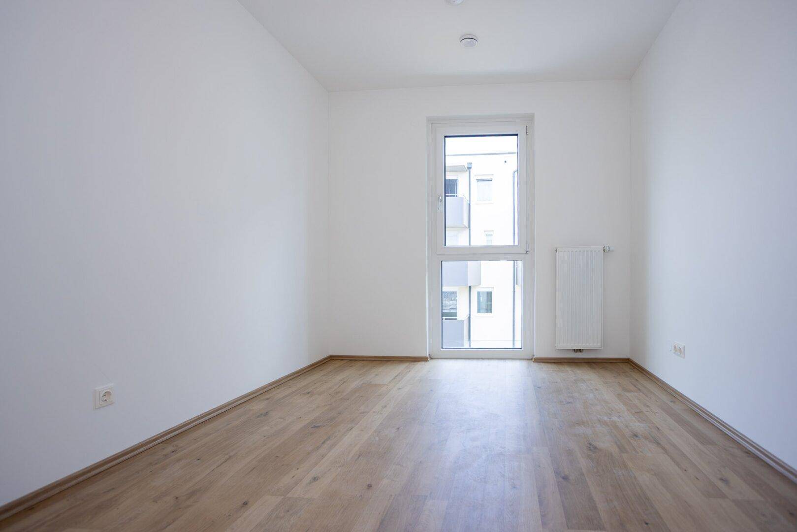 Helles Schlafzimmer mit Laminatboden und einem großen Fenster, das viel Licht hereinlässt.