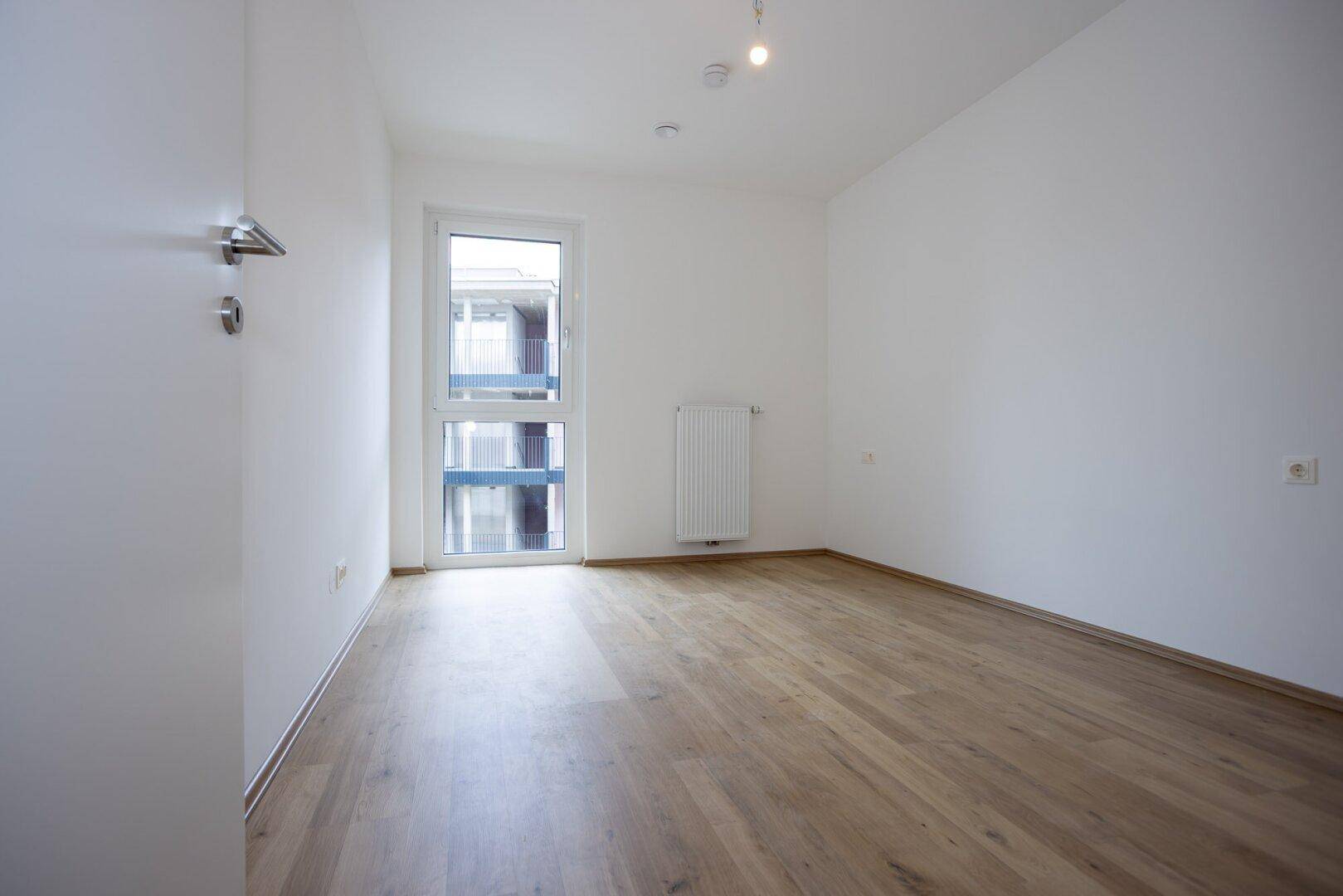 Leeres Zimmer mit hellem Laminatboden und einem Fenster mit Blick auf den Innenhof.