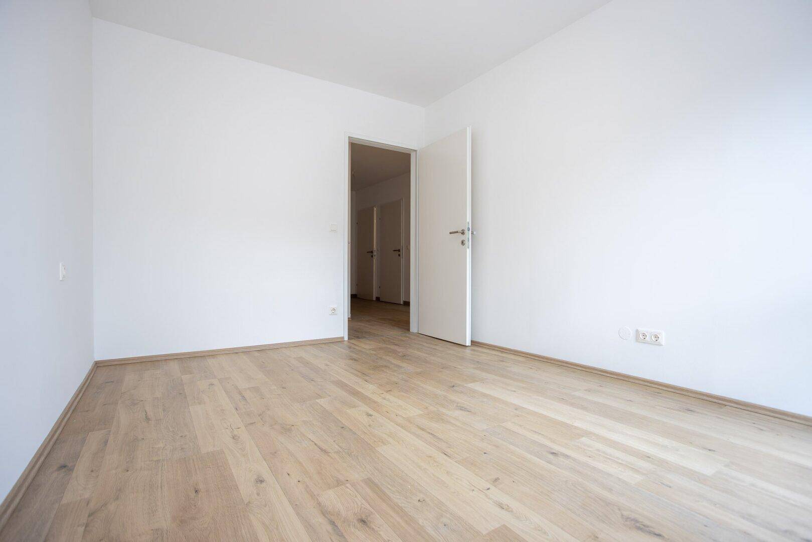 Heller, leerer Raum mit Laminatboden und offener Tür zu einem weiteren Zimmer.