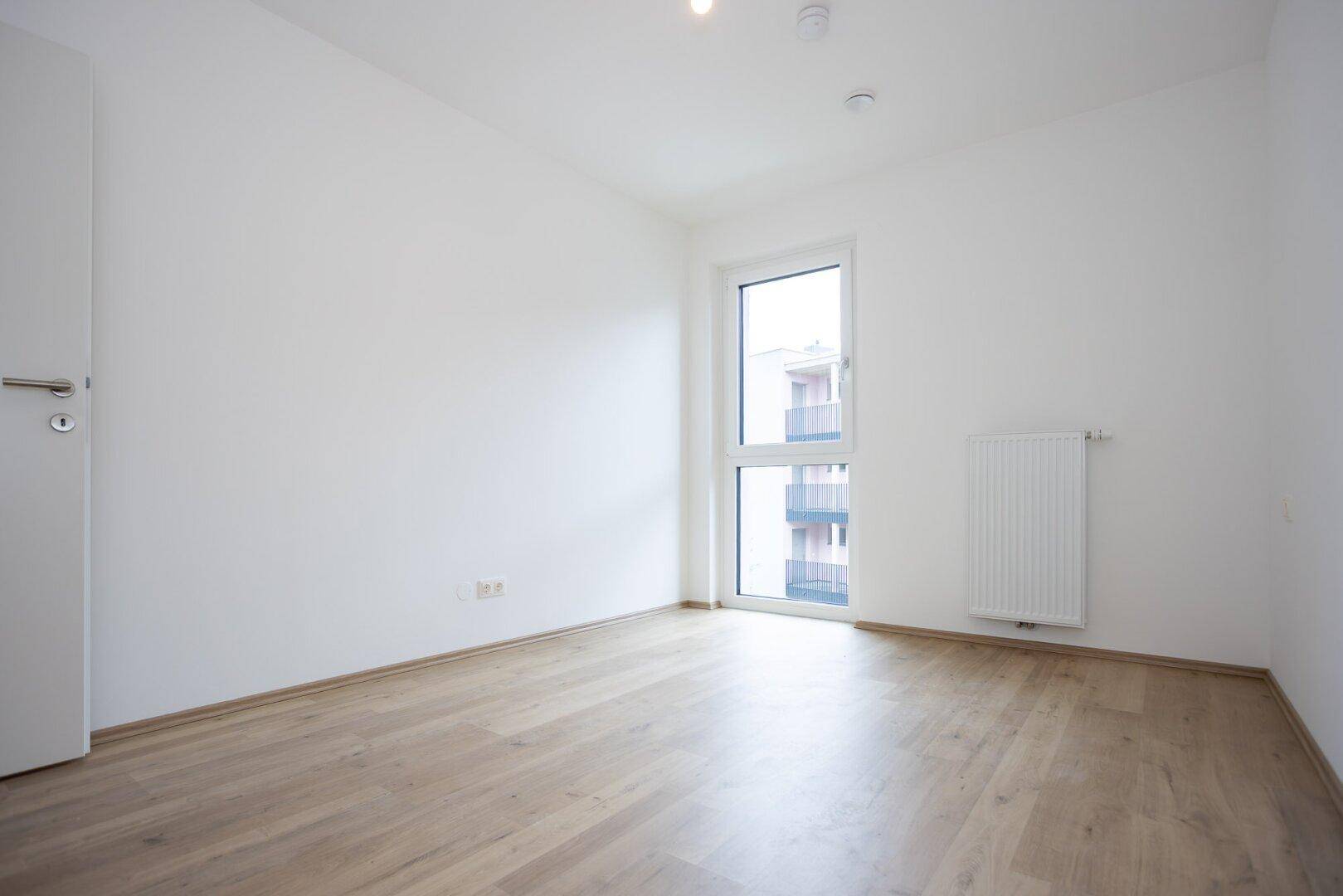 Leeres Zimmer mit hellem Laminatboden und einem Fenster, das viel Tageslicht hereinlässt.