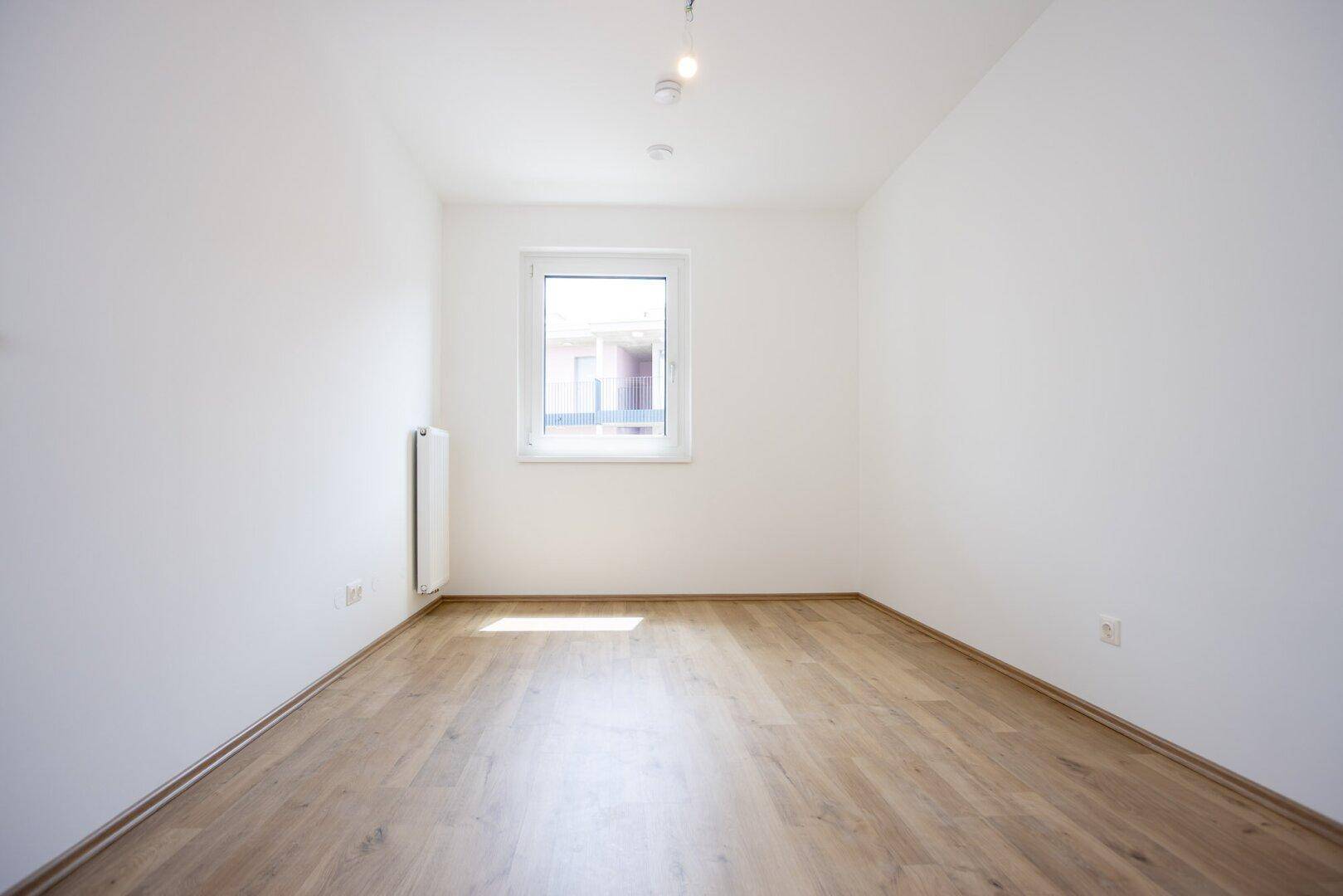 Helles, leeres Zimmer mit Laminatboden und einem einzelnen Fenster.