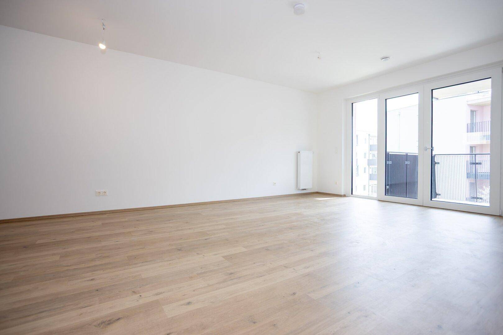Geräumiges, leeres Wohnzimmer mit Laminatboden und großen Fenstern zum Balkon.