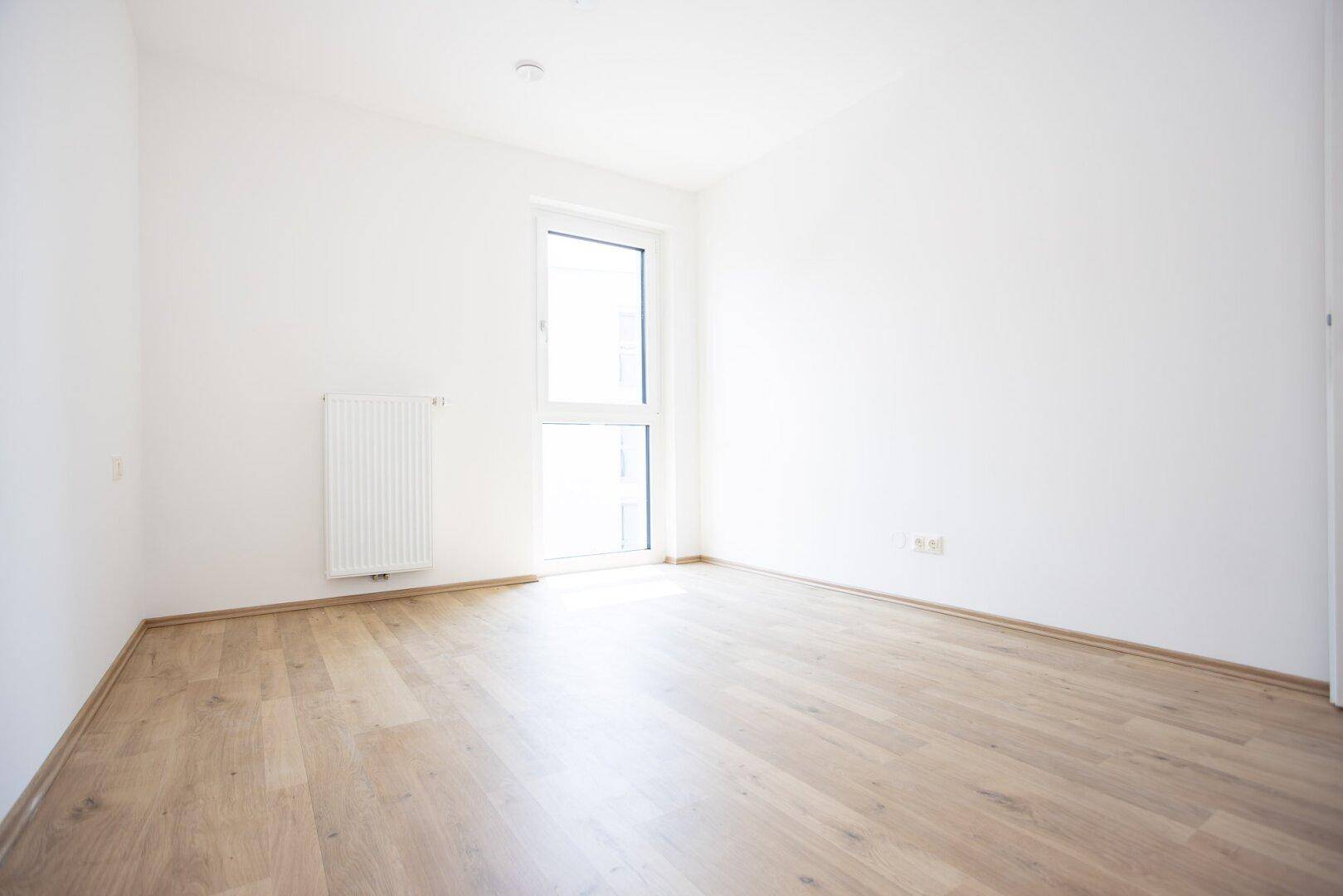 Helles Schlafzimmer mit Parkettboden und einem Fenster, das viel natürliches Licht hereinlässt.