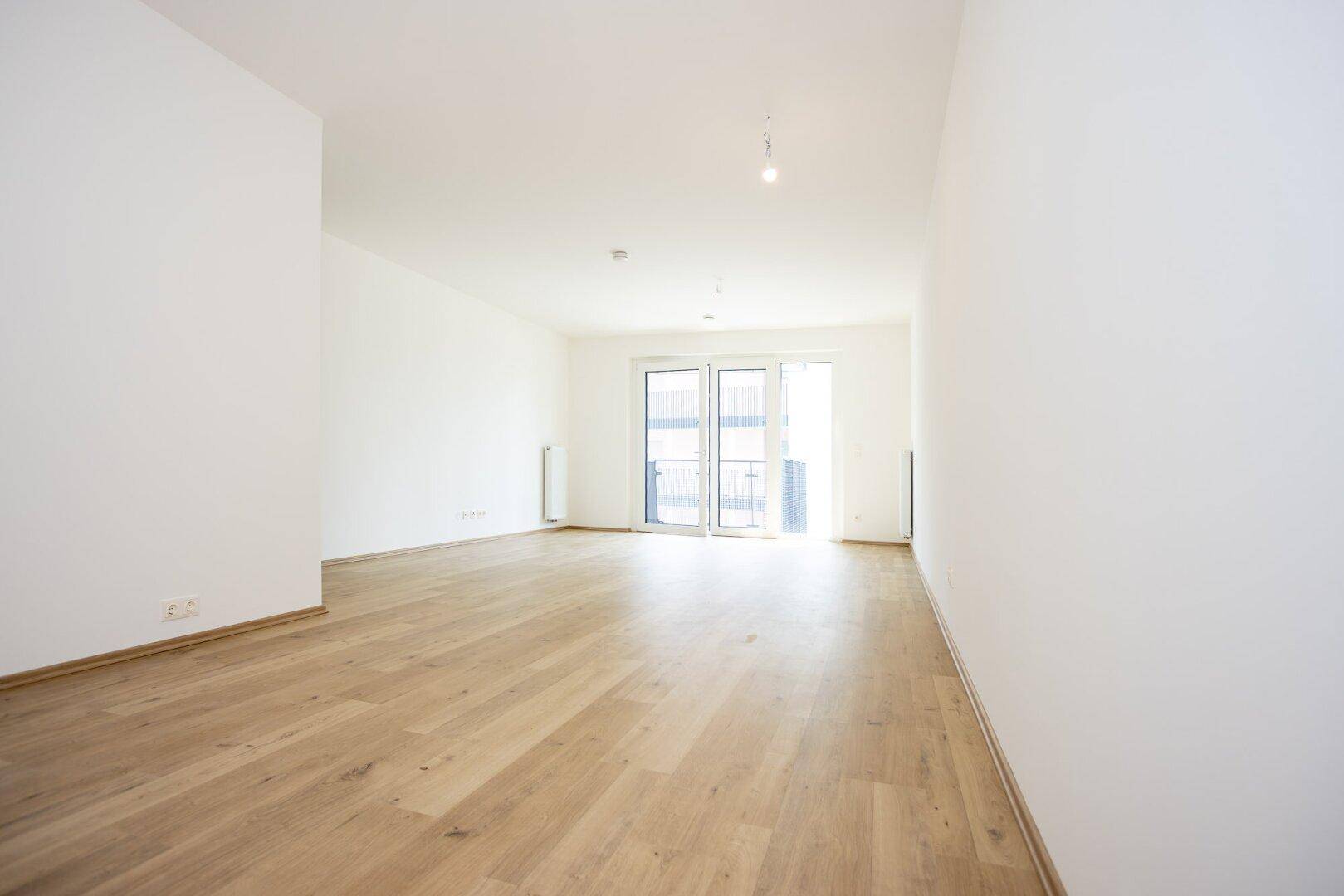 Helles, leeres Zimmer mit hellem Holzboden und Zugang zu einem Balkon.