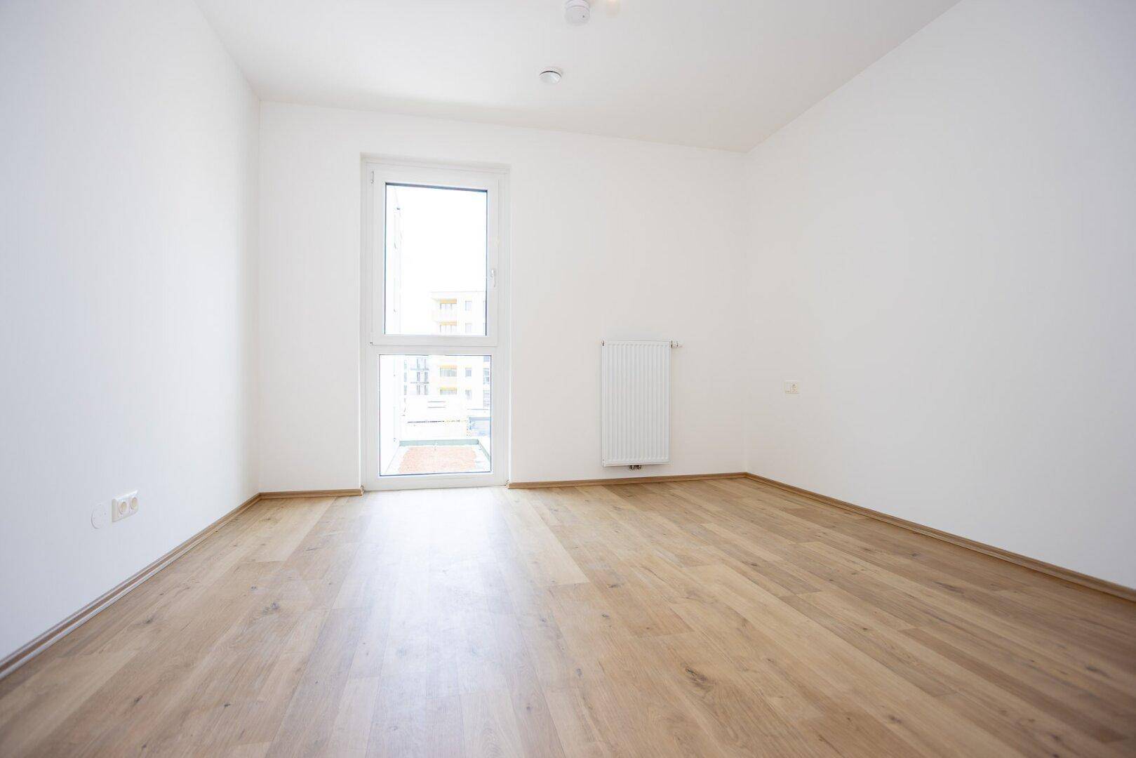 Leeres Zimmer mit hellem Laminatboden, weiß gestrichenen Wänden und einem großen Fenster.