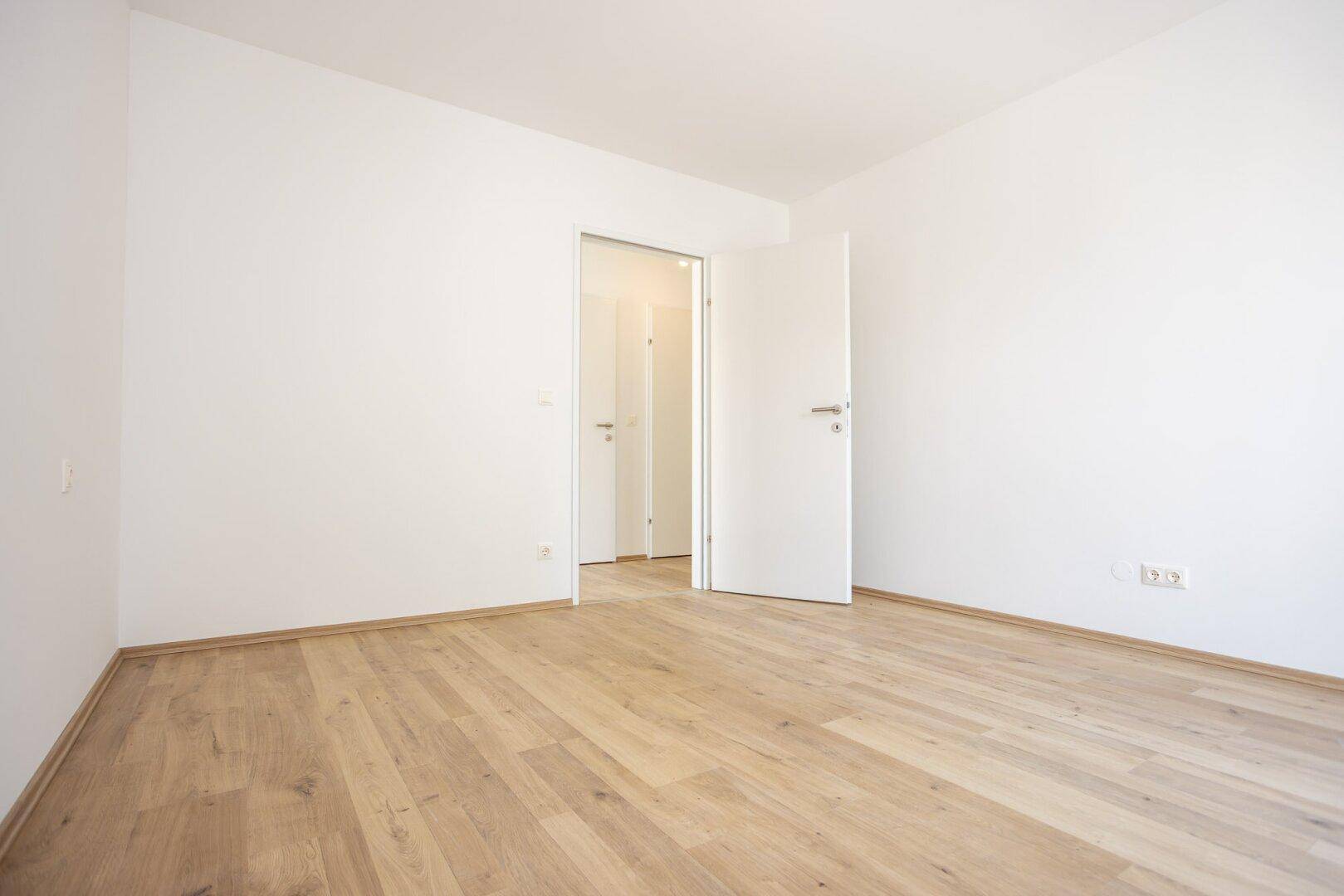 Helles, leeres Zimmer mit Laminatboden und weiß gestrichenen Wänden in einem Neubau.