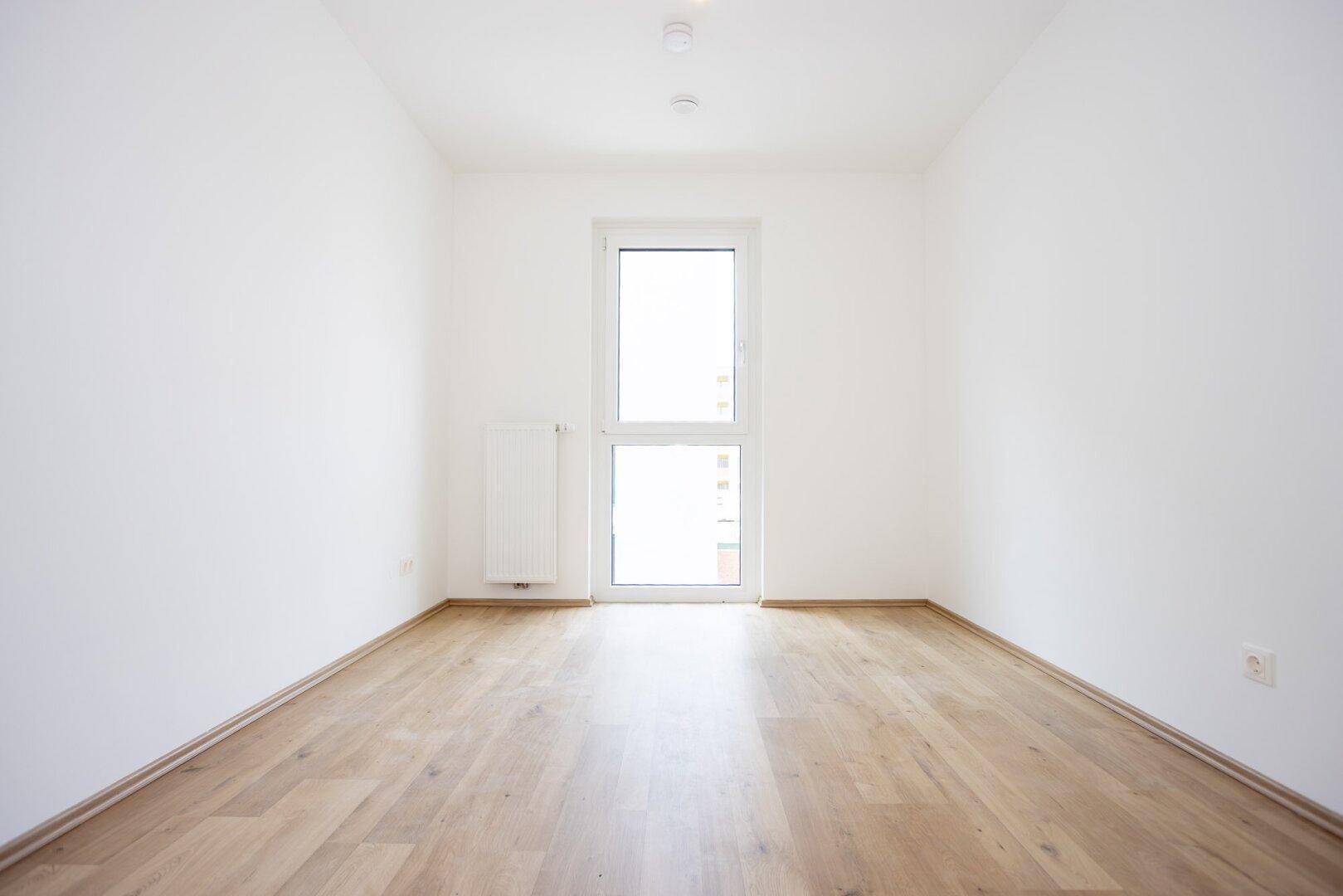 Leeres Zimmer mit hellem Laminatboden, weißen Wänden und einem hohen Fenster.