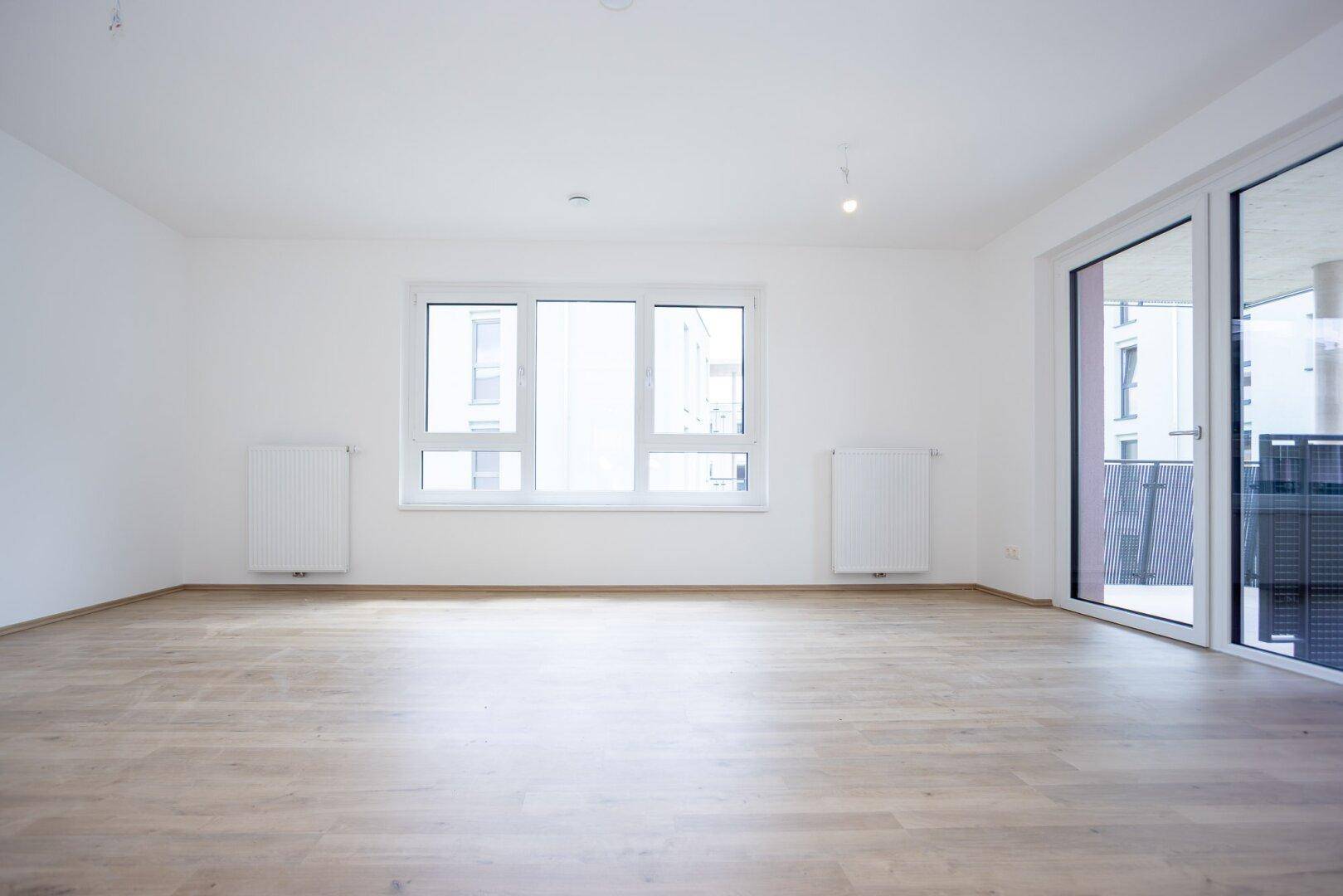Geräumiges Wohnzimmer mit hellem Laminatboden, großen Fenstern und Balkonzugang.