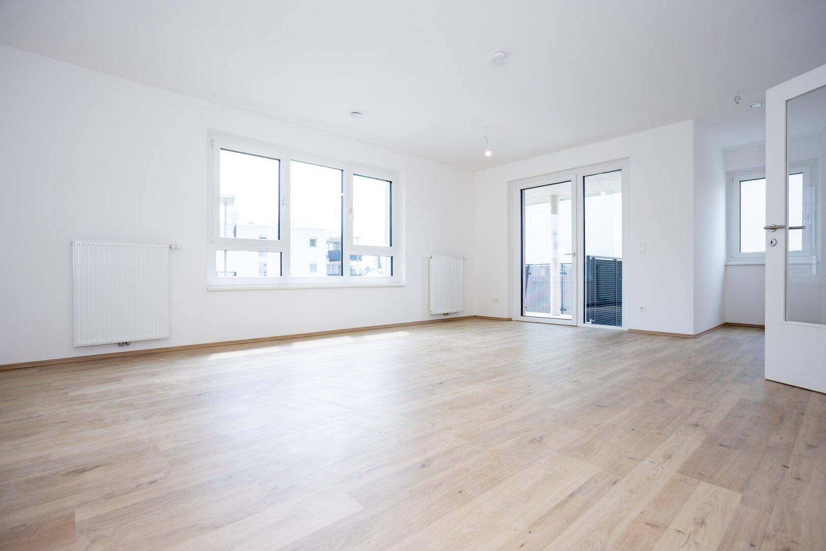 Helles Wohnzimmer mit Laminatboden, großen Fenstern und Zugang zu einem Balkon.