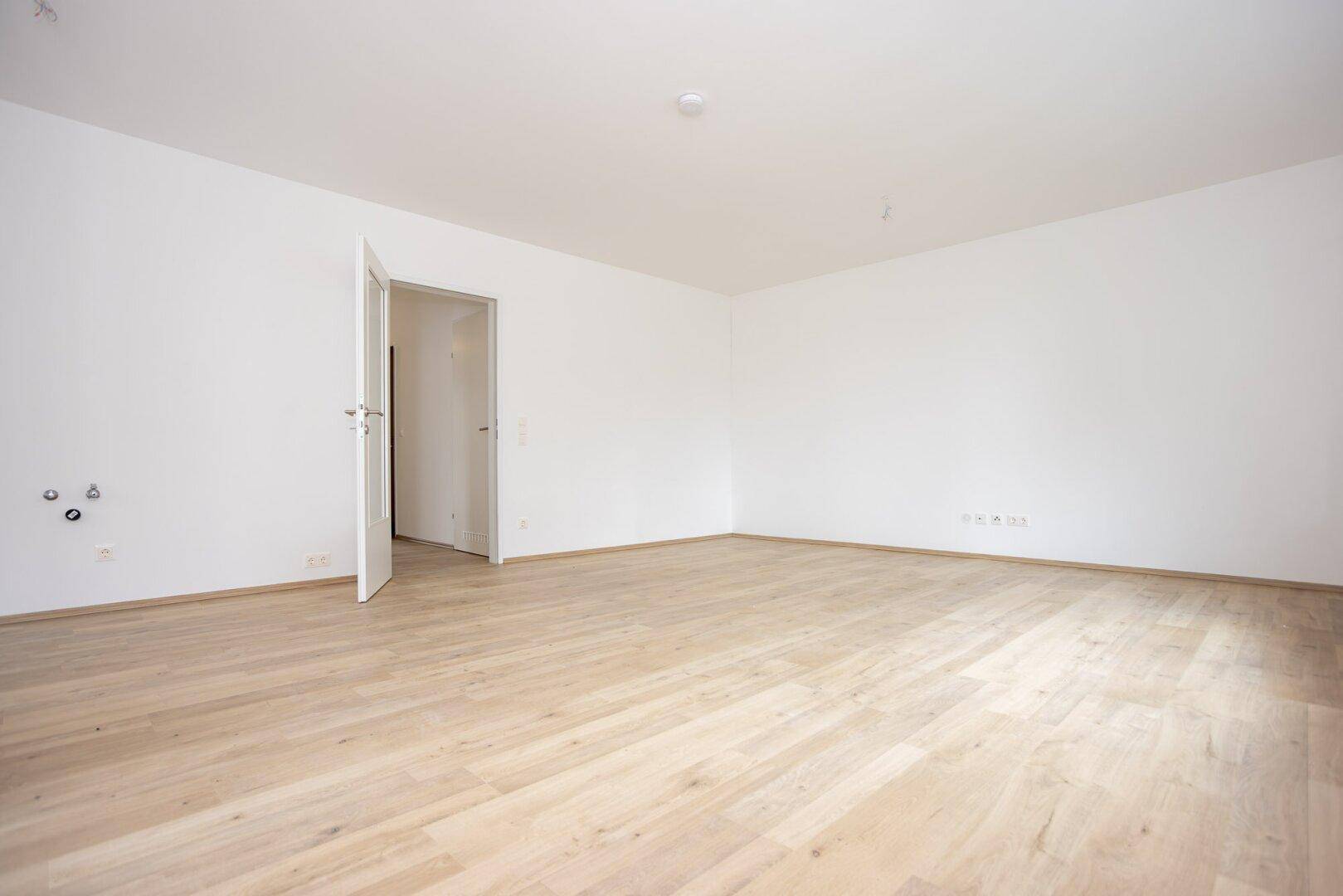 Geräumiges, leeres Zimmer mit hellem Laminatboden und weiß gestrichenen Wänden.