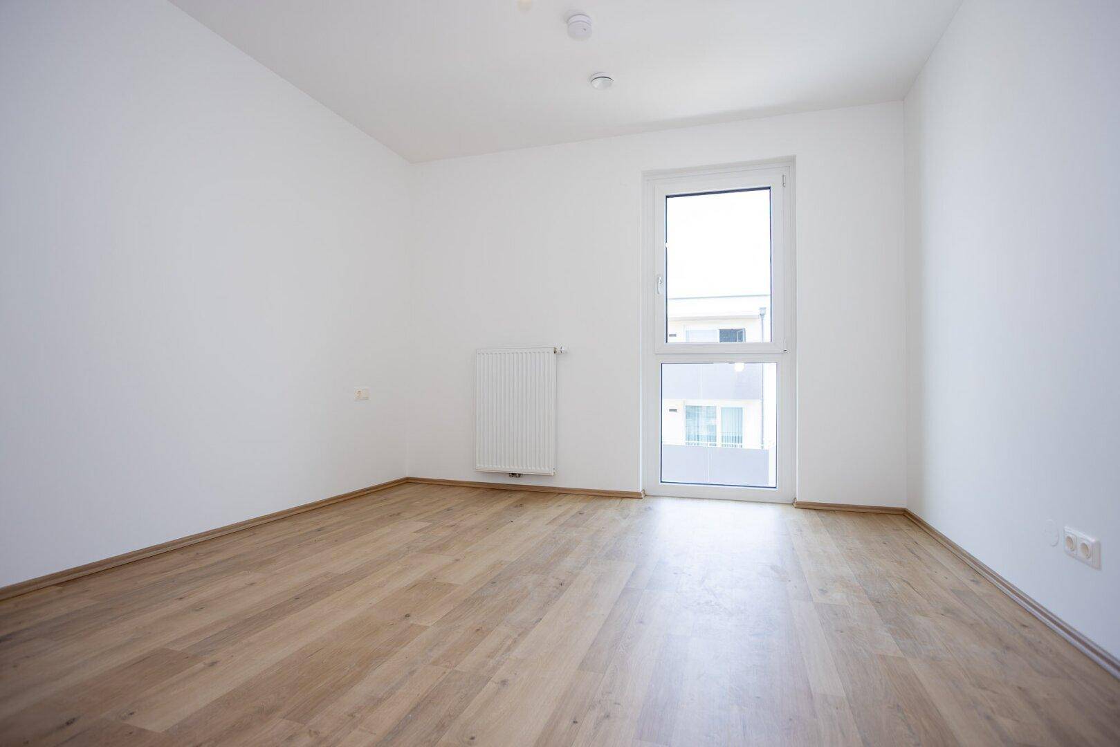 Helles Schlafzimmer mit Laminatboden und einem großen Fenster, das viel Tageslicht hereinlässt.