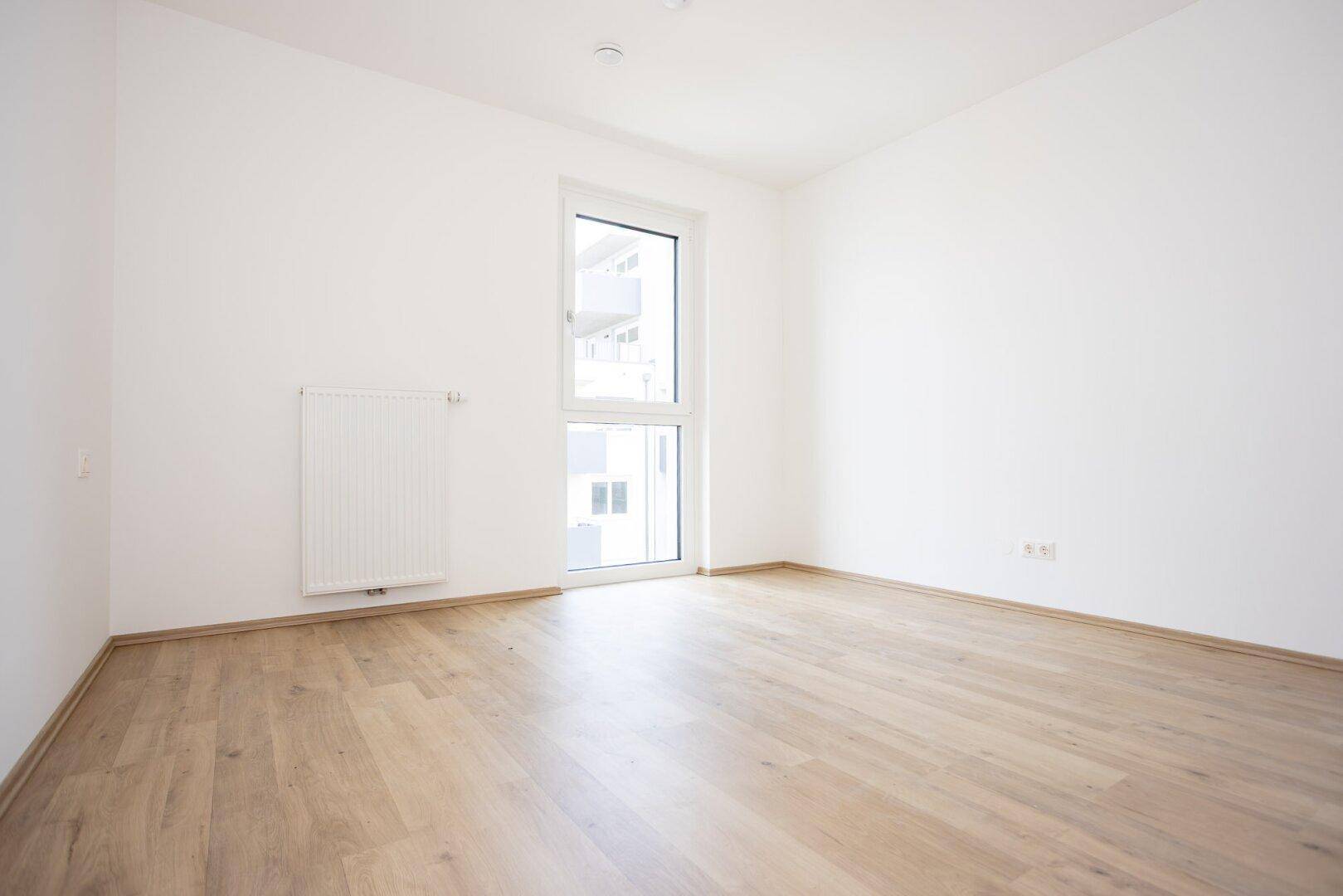 Geräumiges Zimmer mit hellem Laminatboden und einem hohen Fenster, ideal als Schlaf- oder Arbeitszimmer.