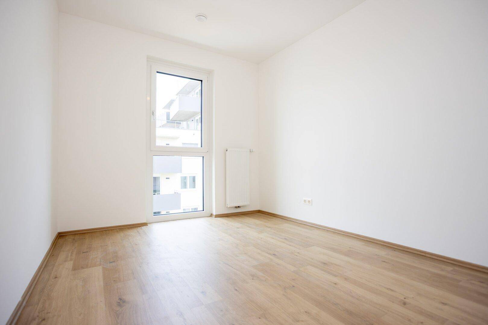 Freundliches Zimmer mit hellem Laminatboden und einem großen Fenster, das eine angenehme Atmosphäre schafft.