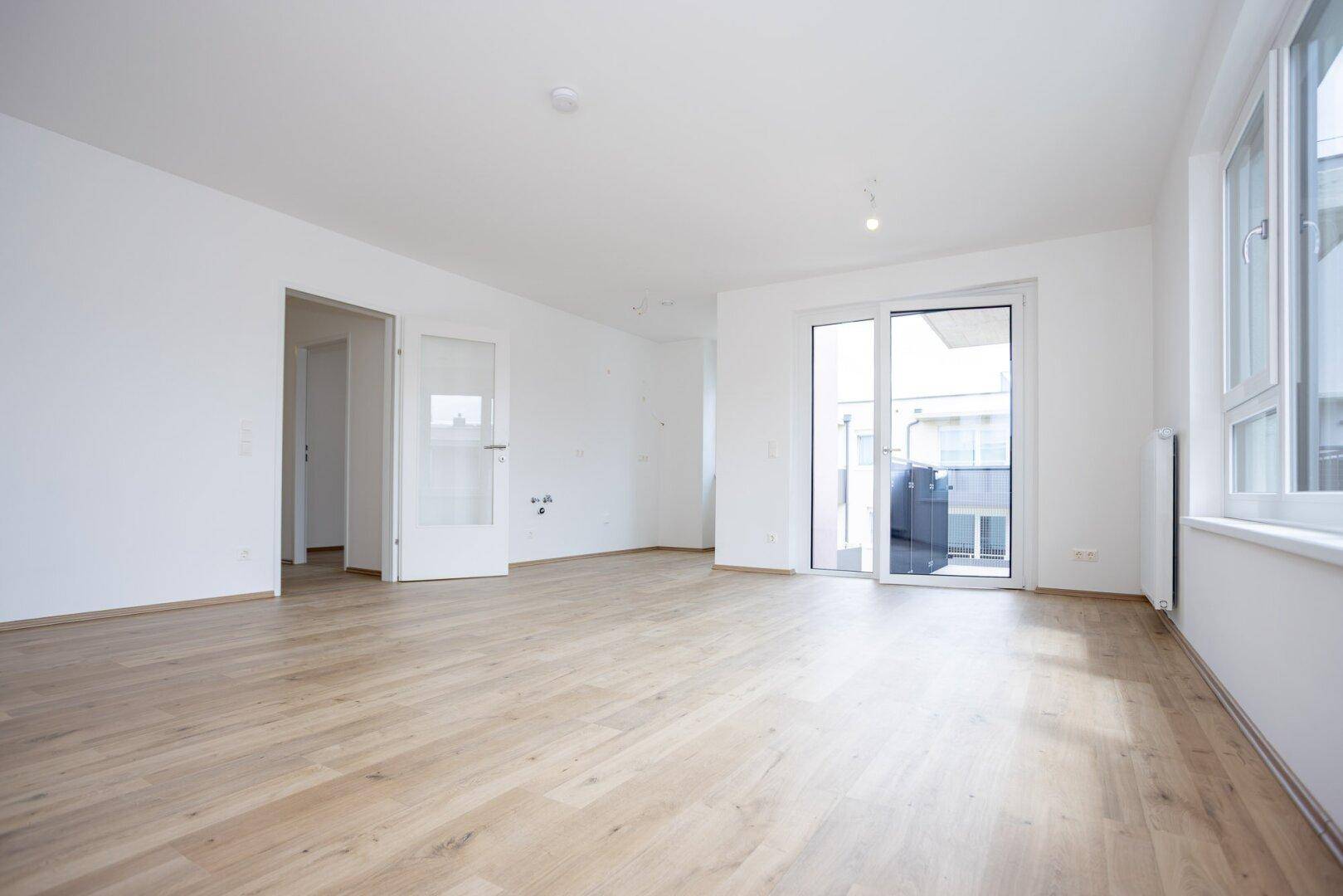 Geräumiges Wohnzimmer mit hellem Laminatboden, großen Fenstern und Balkonzugang.
