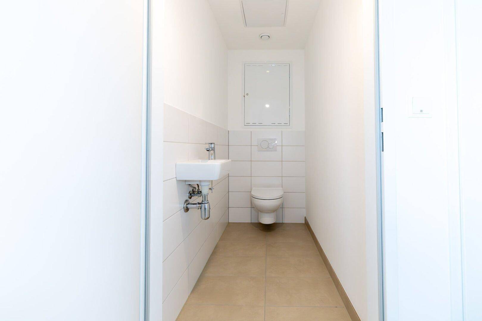 Modernes Gäste-WC mit Wand-WC, kleinem Waschbecken und hellen Fliesen, funktional gestaltet.
