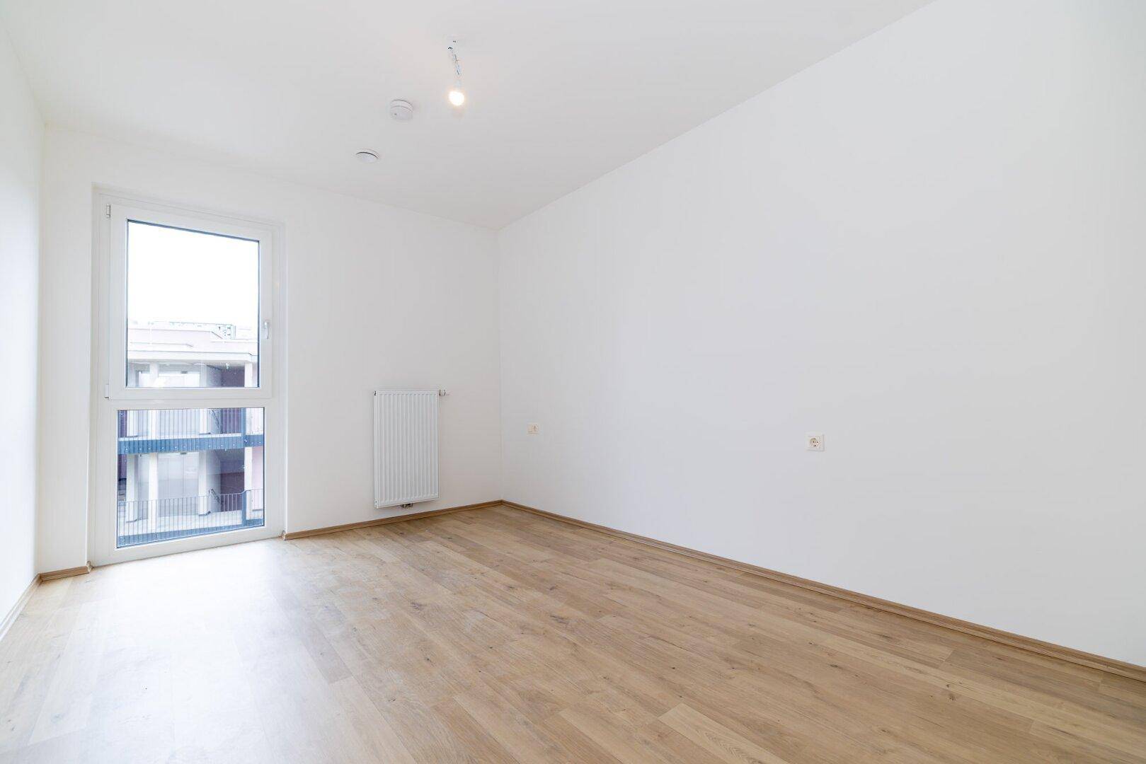 Schlafzimmer mit Laminatboden und einem bodentiefen Fenster, das viel Licht hereinlässt.