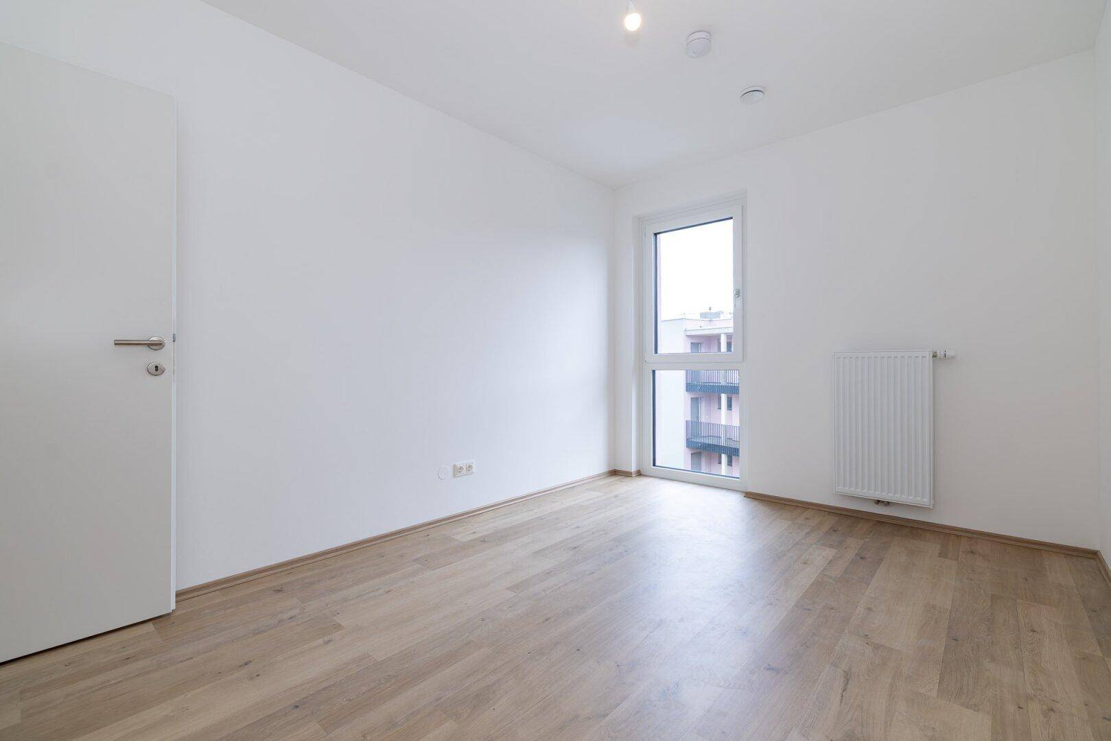 Helles Schlafzimmer mit Laminatboden und einem Fenster, das Zugang zu einem kleinen Balkon bietet.