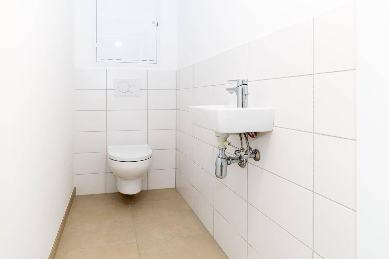 Separates WC mit wandhängender Toilette, kleinem Waschbecken und weißen Fliesen.