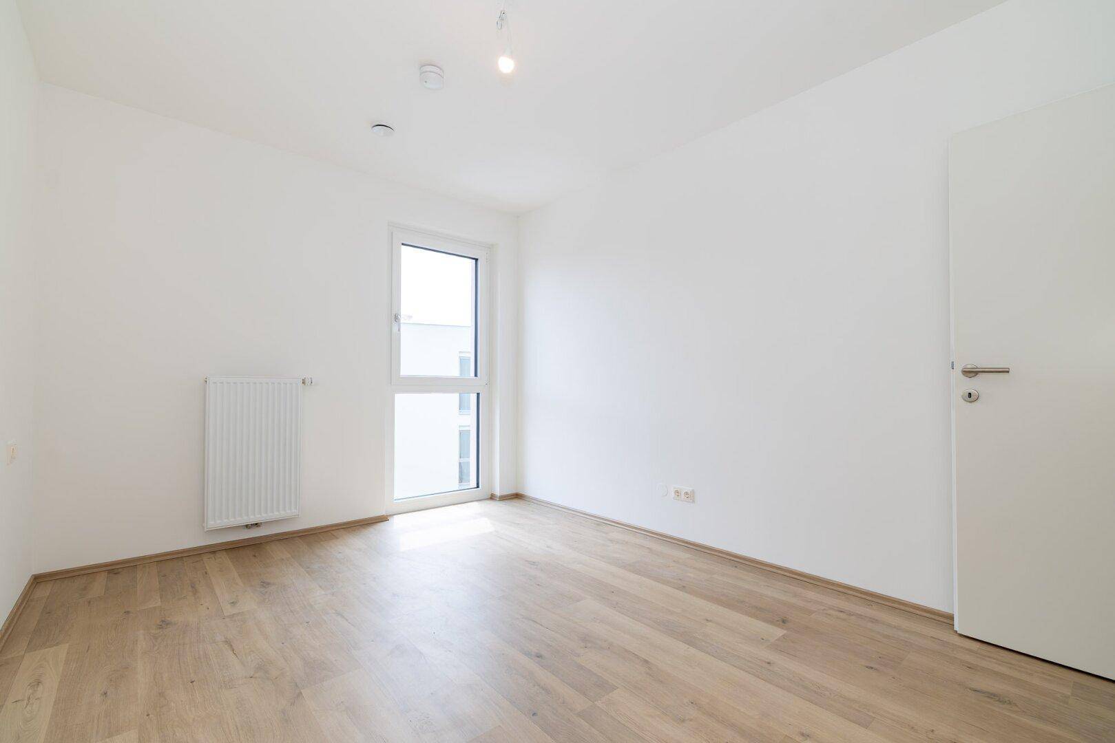 Leeres Schlafzimmer mit hellem Laminatboden, weißen Wänden und einem großen Fenster.