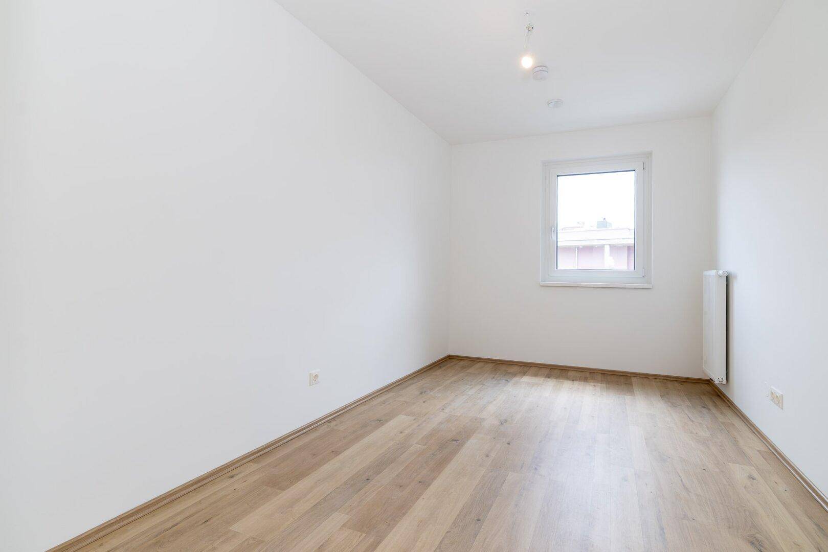 Leeres Schlafzimmer mit hellem Laminatboden, weißen Wänden und einem Fenster.