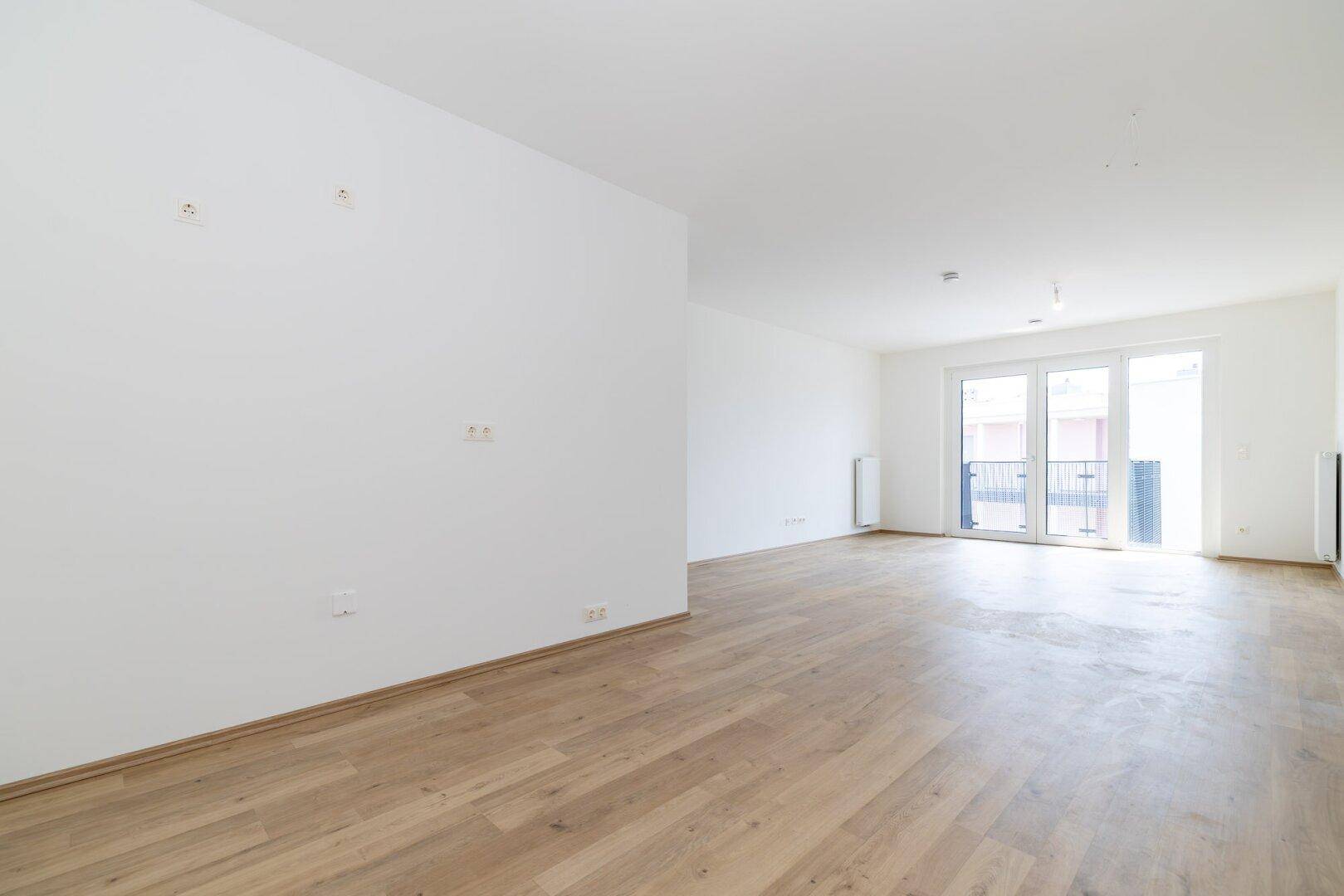 Heller, leerer Wohnraum mit modernem Laminatboden, weißen Wänden und Zugang zum Balkon.