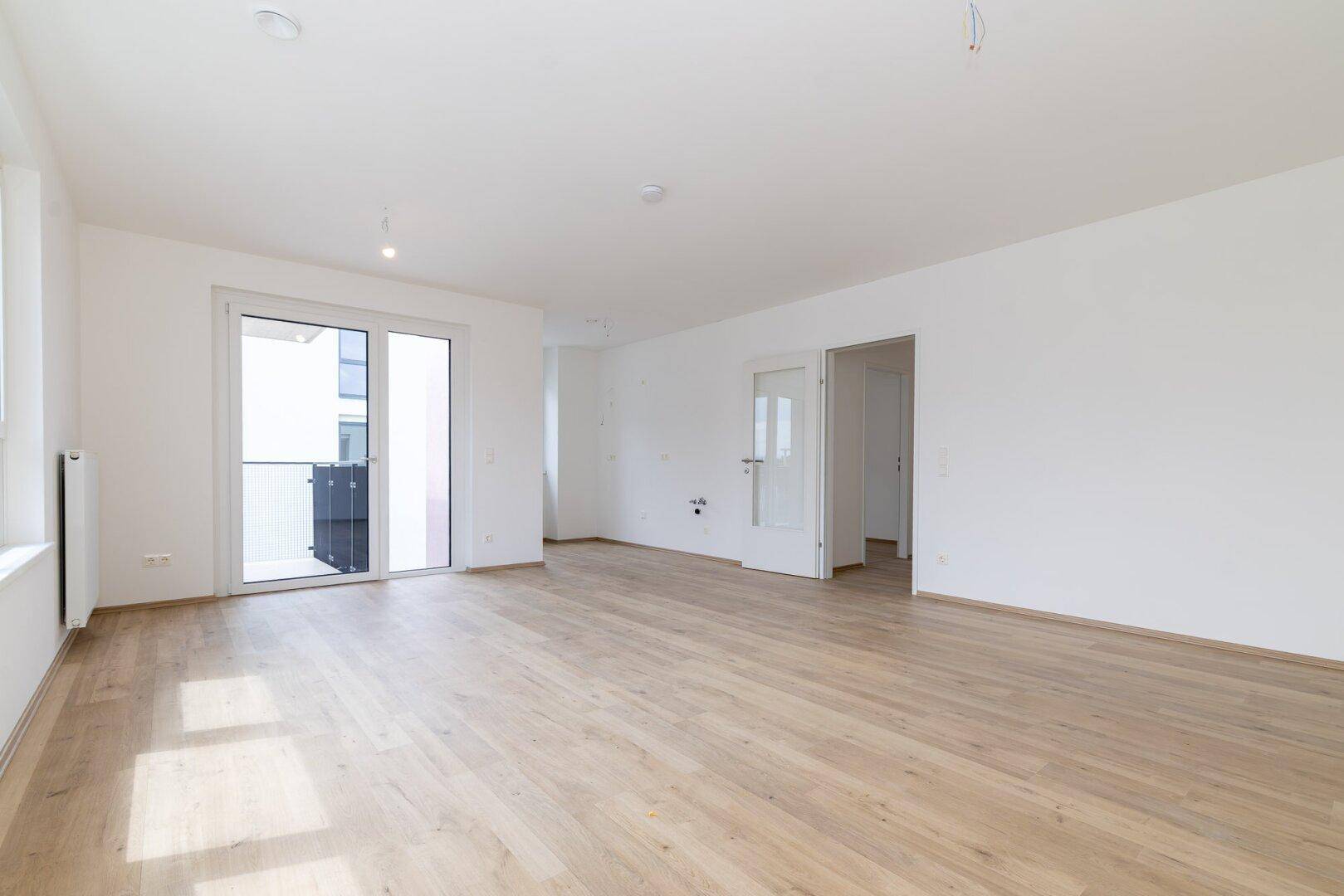 Heller Wohnbereich mit Holzboden, großen Fenstern und Zugang zum Balkon.