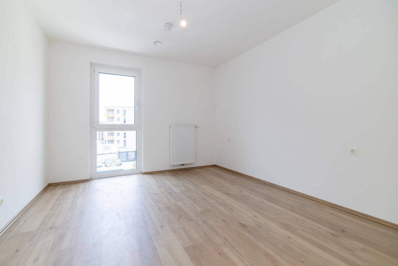 Helles Schlafzimmer mit Holzboden und einem hohen Fenster, das viel Licht hereinlässt.