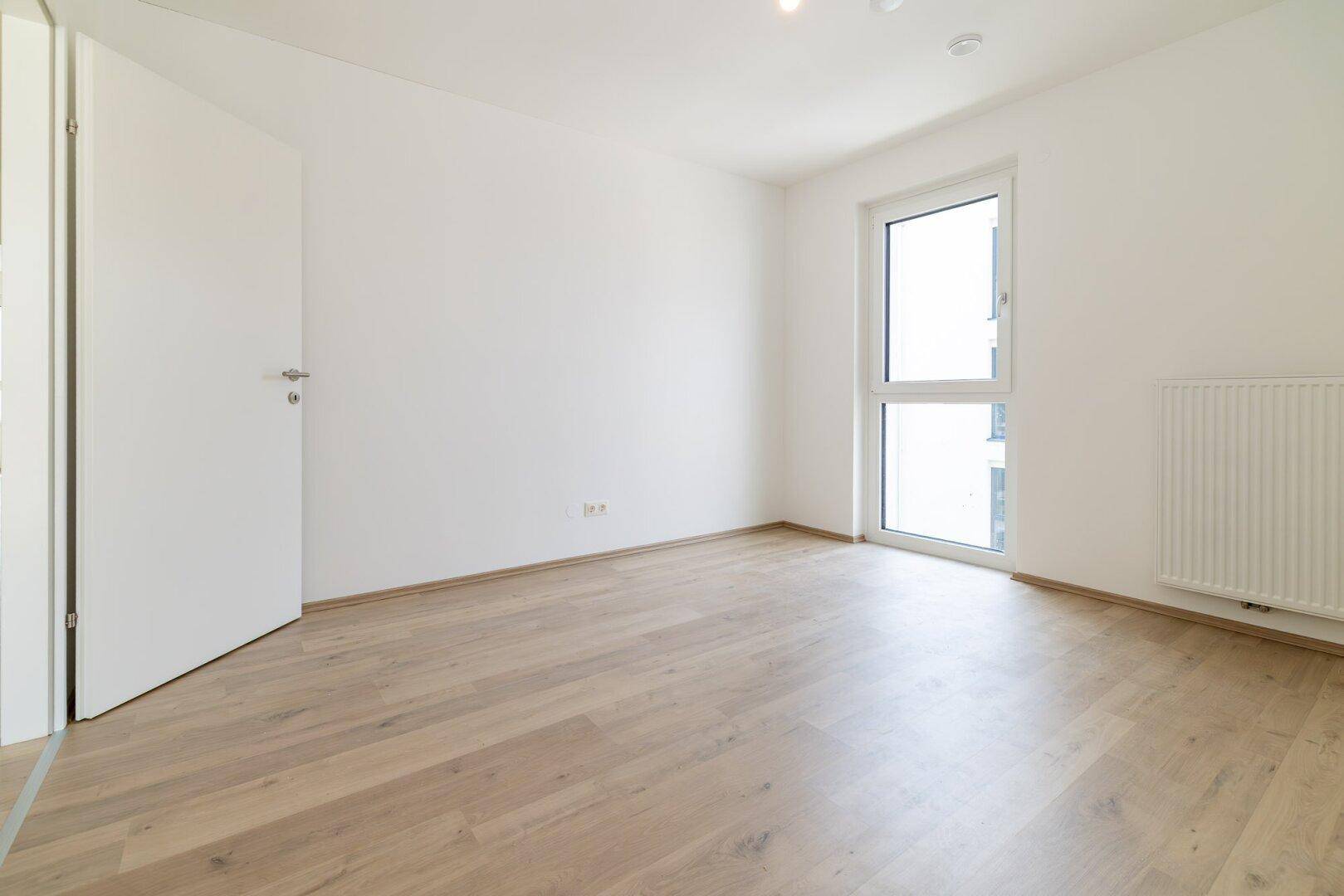 Leeres Schlafzimmer mit hellem Holzboden und einem großen Fenster für viel Tageslicht.