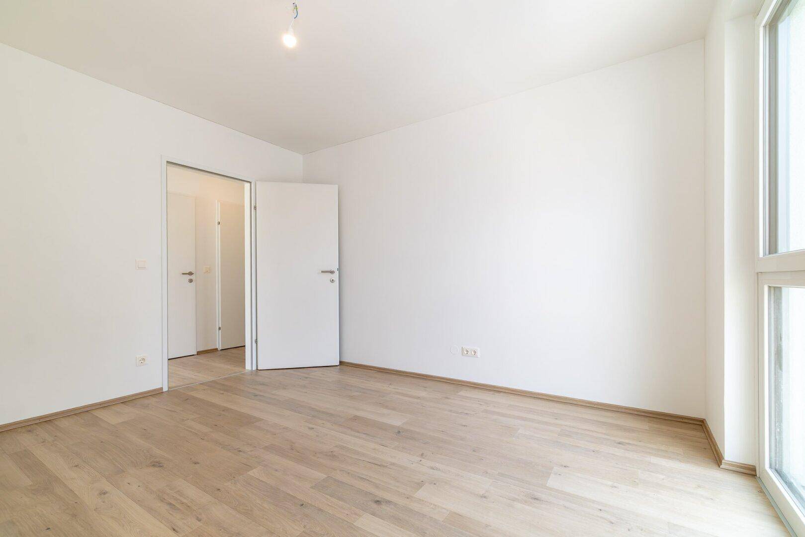 Leerer Raum mit hellem Holzboden und offener Tür zu einem weiteren Zimmer.