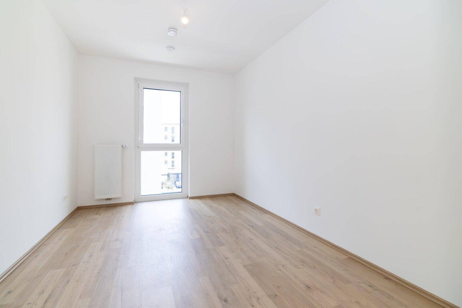 Helles Schlafzimmer mit Holzboden und einem hohen Fenster, das den Raum erhellt.