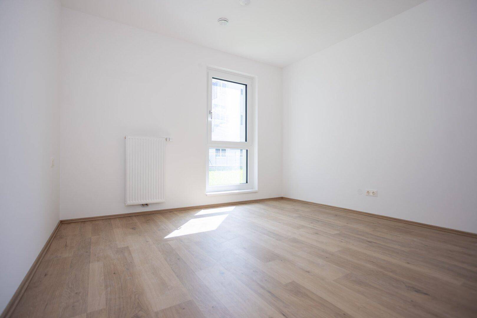 Leeres Schlafzimmer mit Laminatboden, weiß gestrichenen Wänden und einem hohen Fenster.