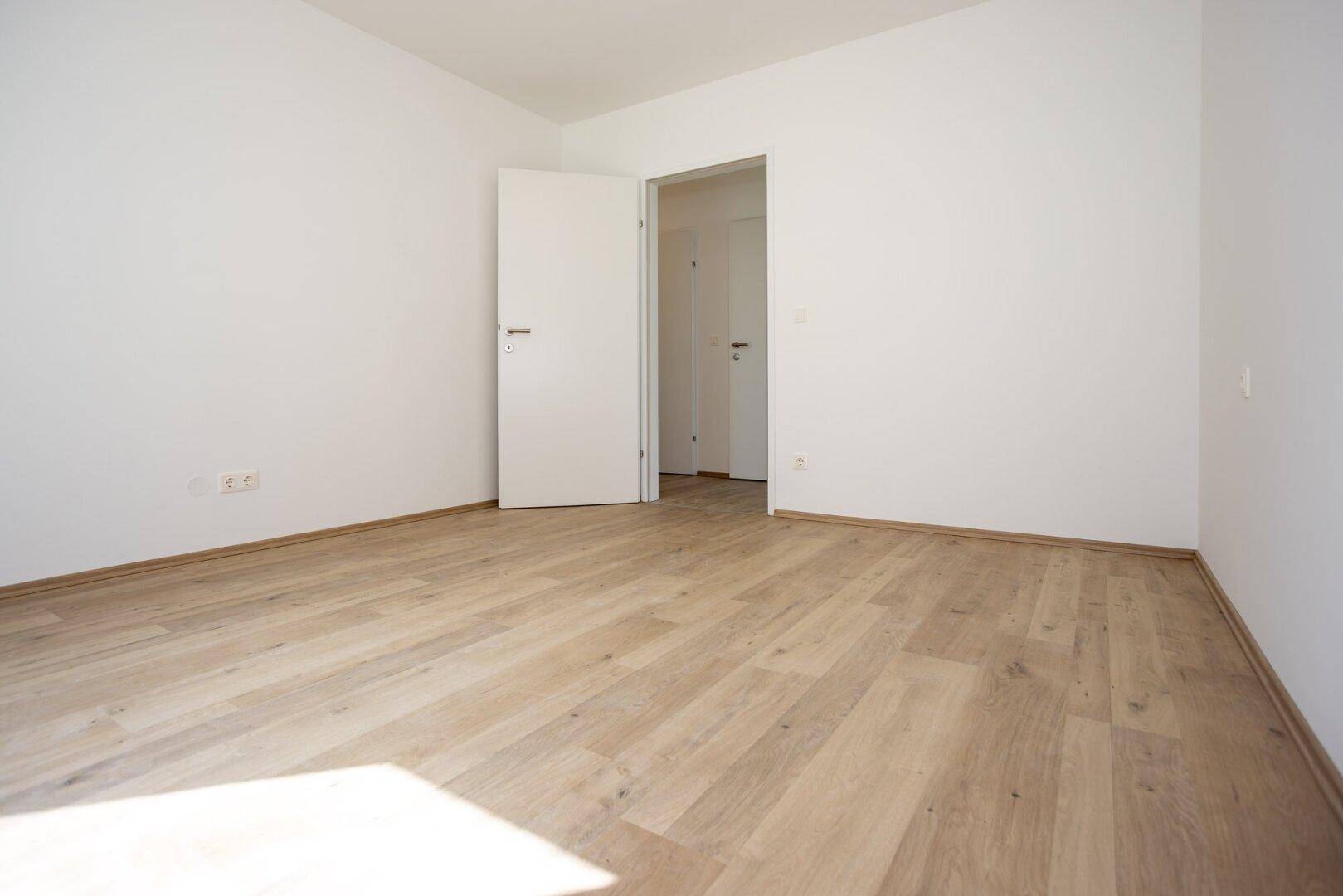Leeres Zimmer mit Laminatboden, weißen Wänden und einer offenen Tür.
