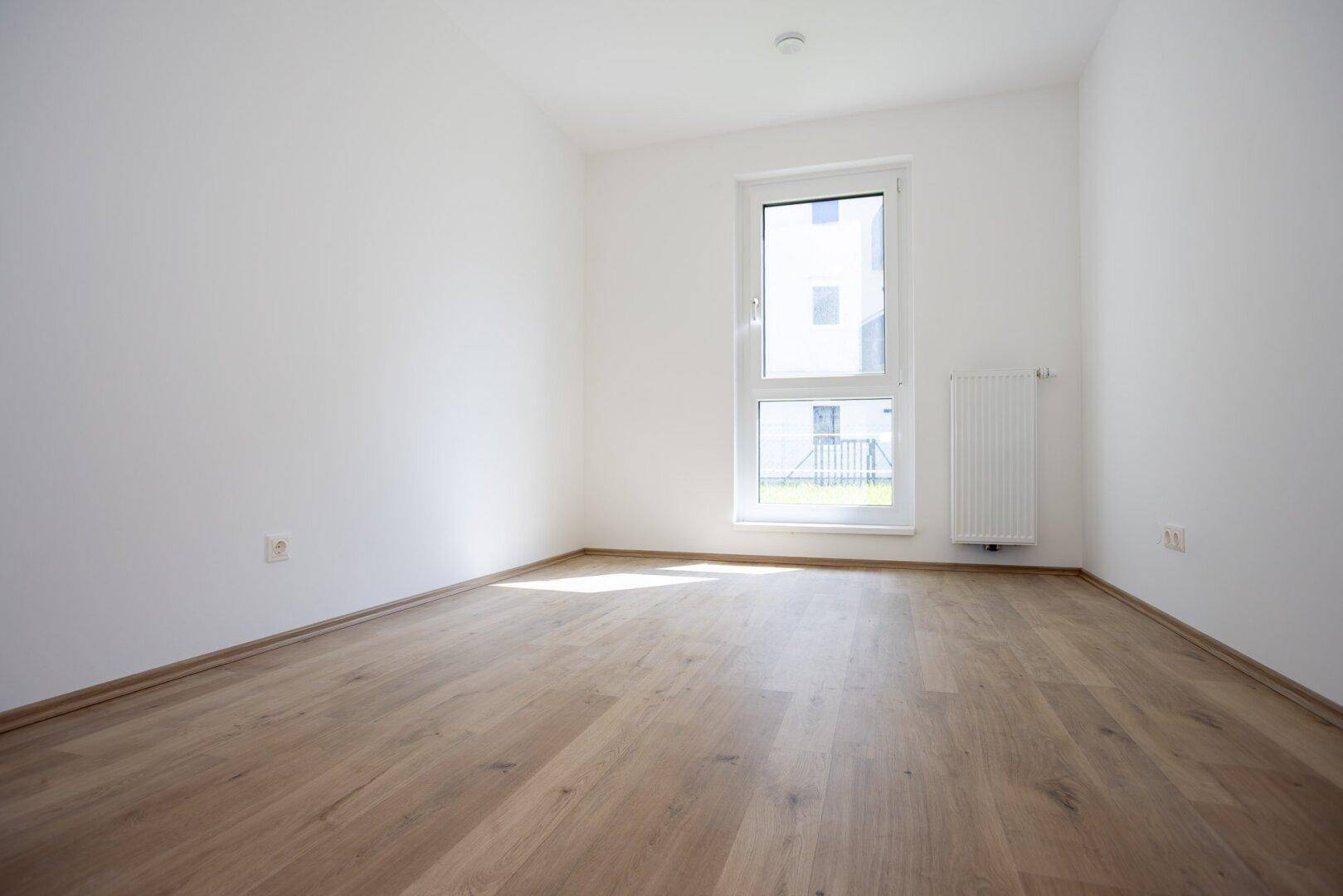 Helles, leeres Zimmer mit Laminatboden und einem Fenster mit Blick ins Grüne.