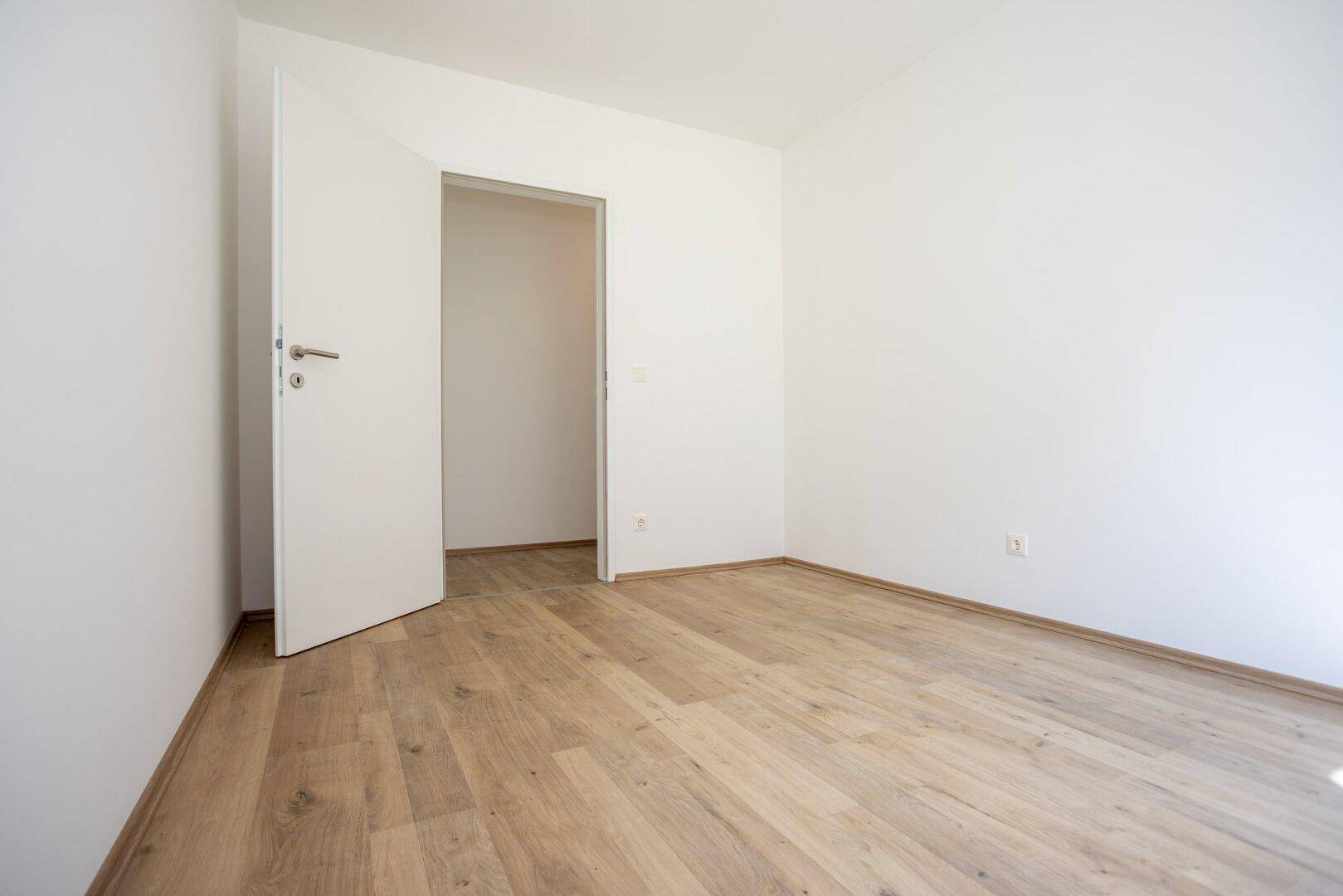 Leerer Raum mit Laminatboden und einer offenen Tür, die in einen weiteren Bereich führt.