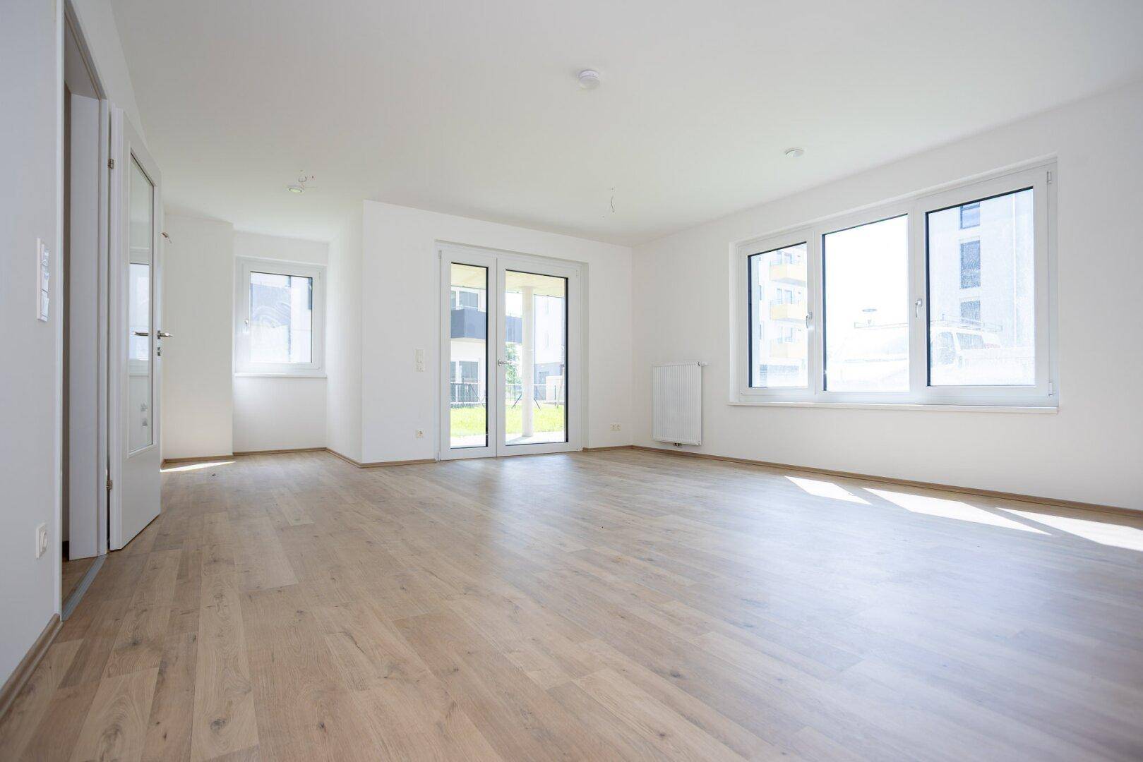 Helles, leeres Wohnzimmer mit großen Fenstern, Laminatboden und Zugang zum Garten.