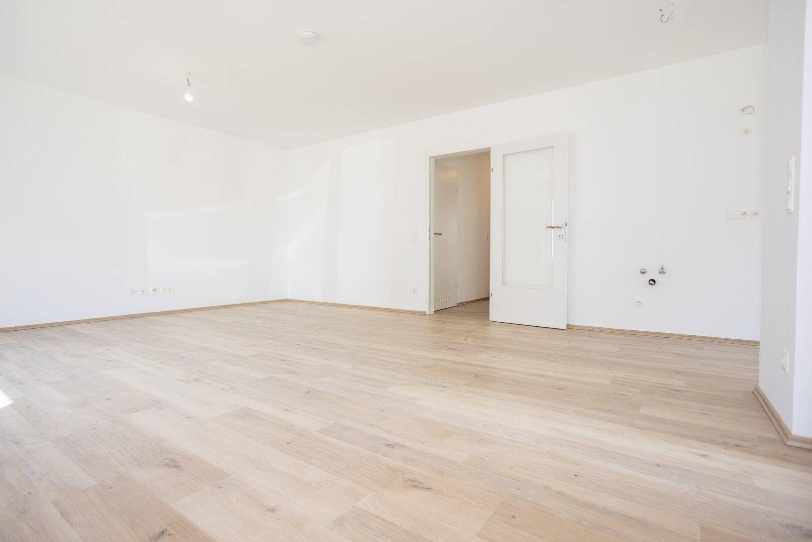 Geräumiger, heller Raum mit Laminatboden und Anschlüssen für eine zukünftige Küche.