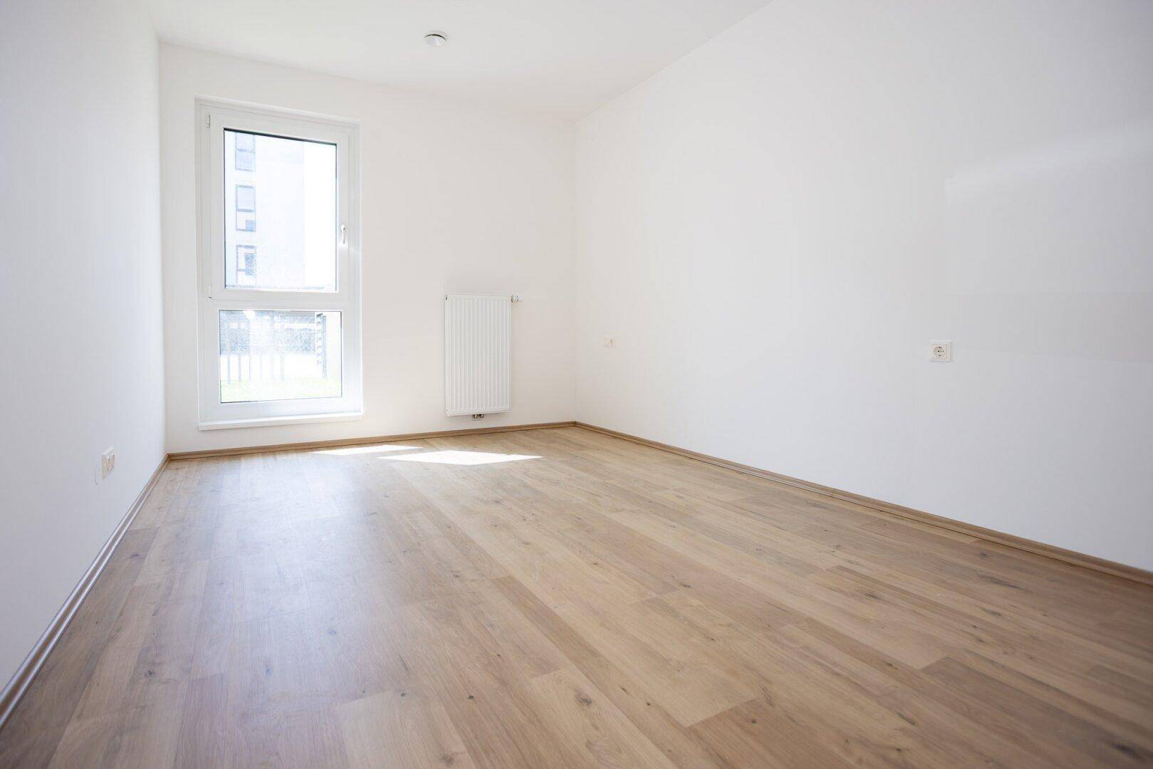 Leeres Schlafzimmer mit hellem Laminatboden und großem Fenster, das viel Licht hereinlässt.