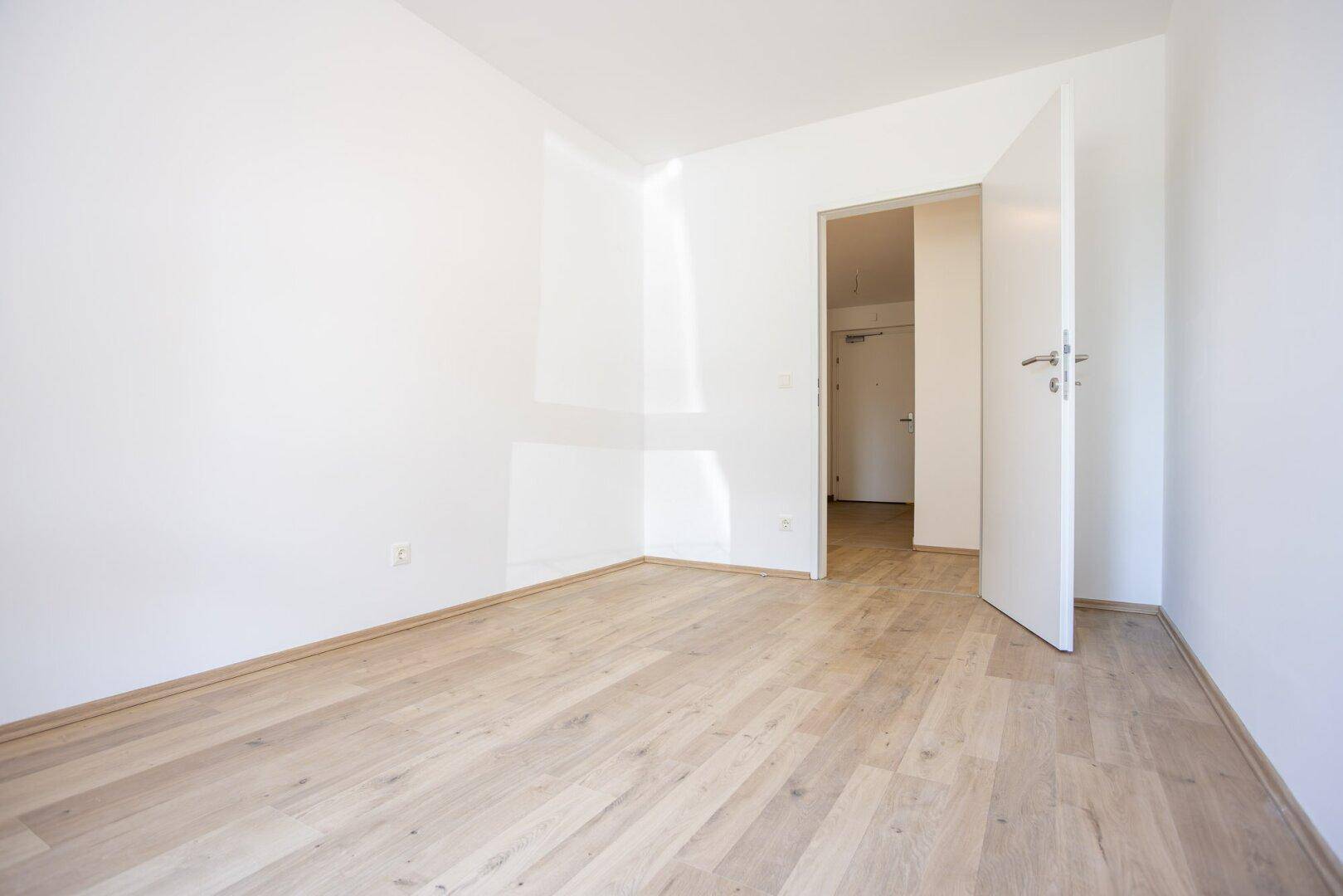 Leeres Zimmer mit Laminatboden und offener Tür zum Flur, in einem modernen Neubau.