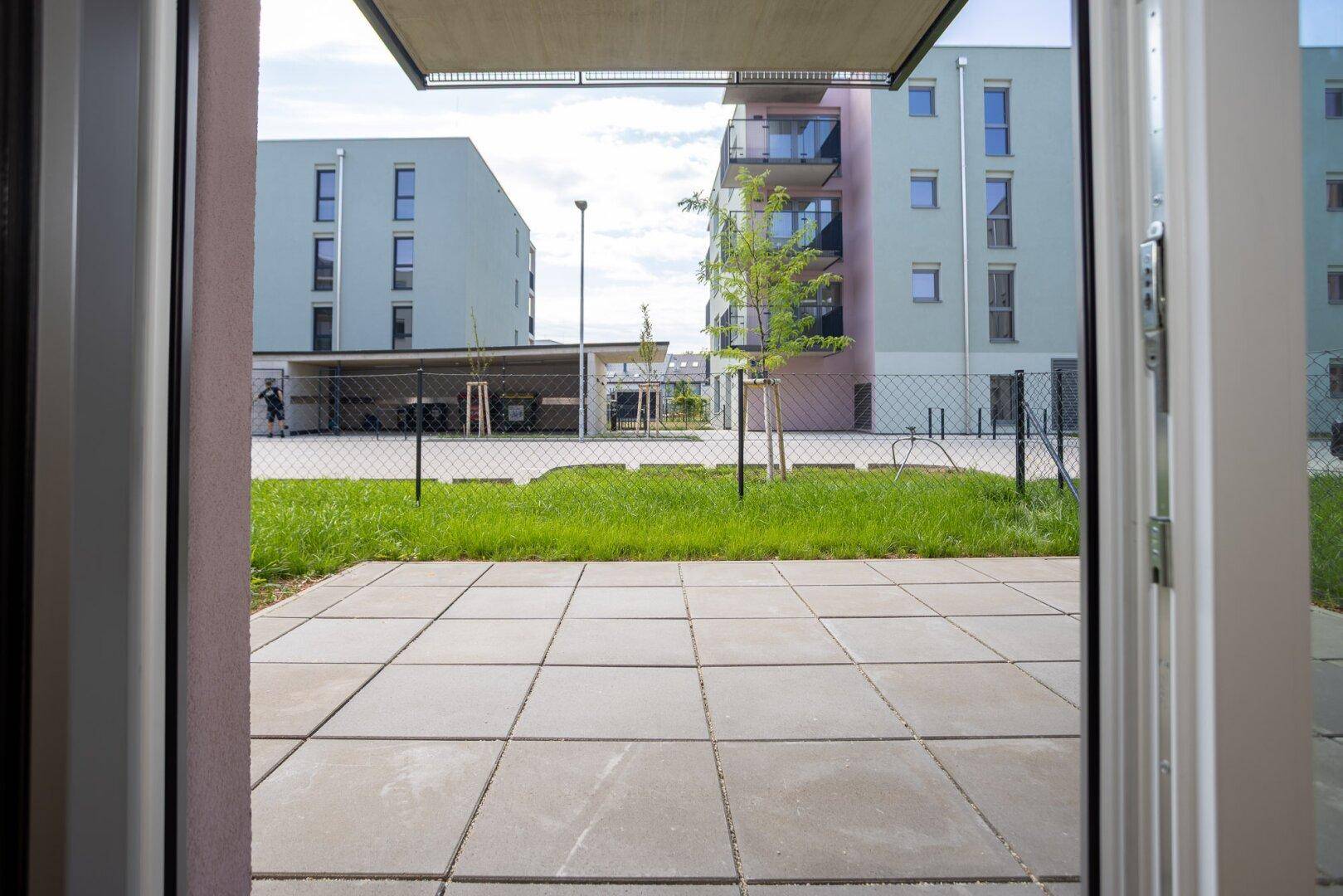 Blick von innen auf eine gepflasterte Terrasse mit Rasenfläche und modernen Wohngebäuden.