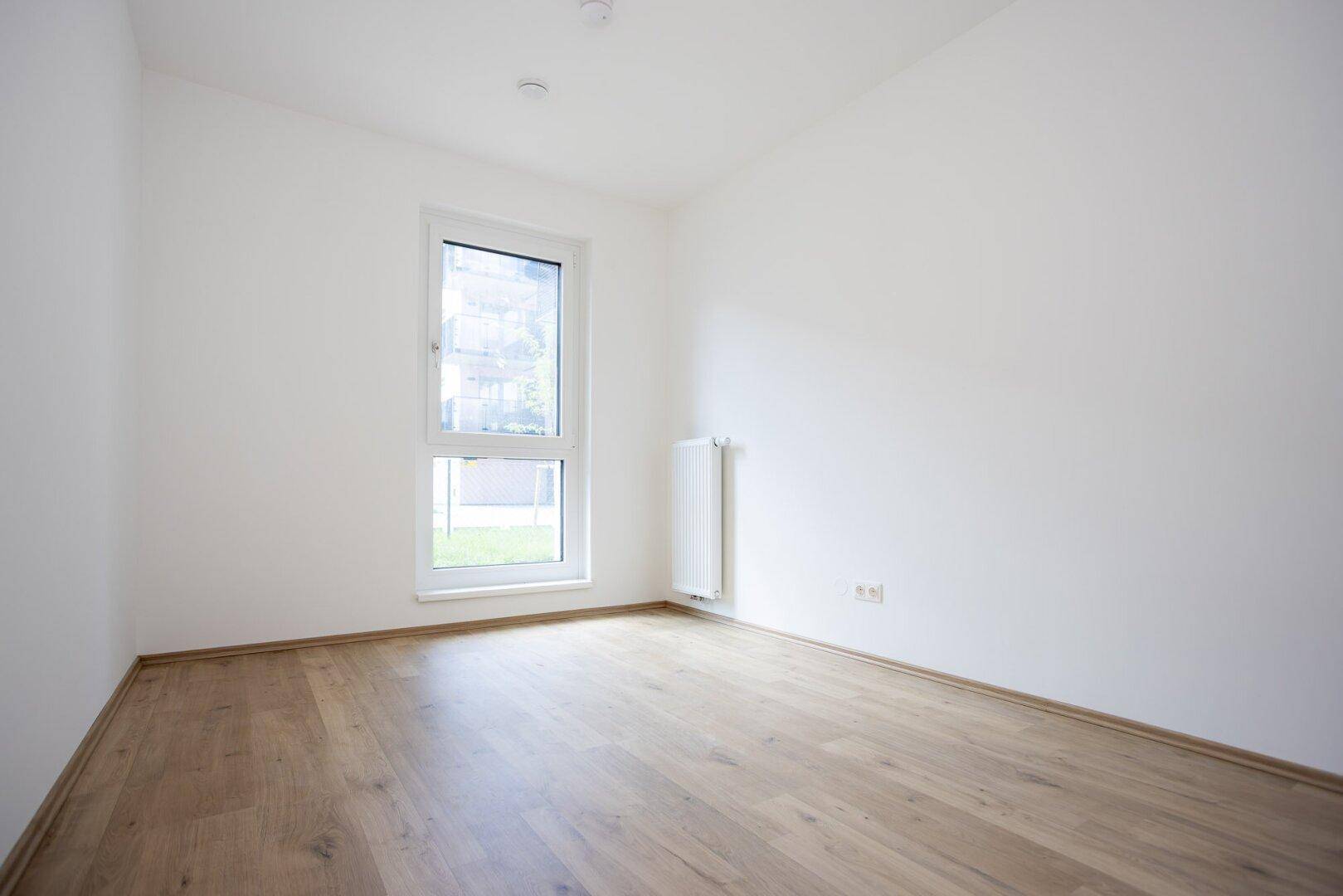 Helles Zimmer mit Laminatboden und einem Fenster mit Blick ins Grüne.
