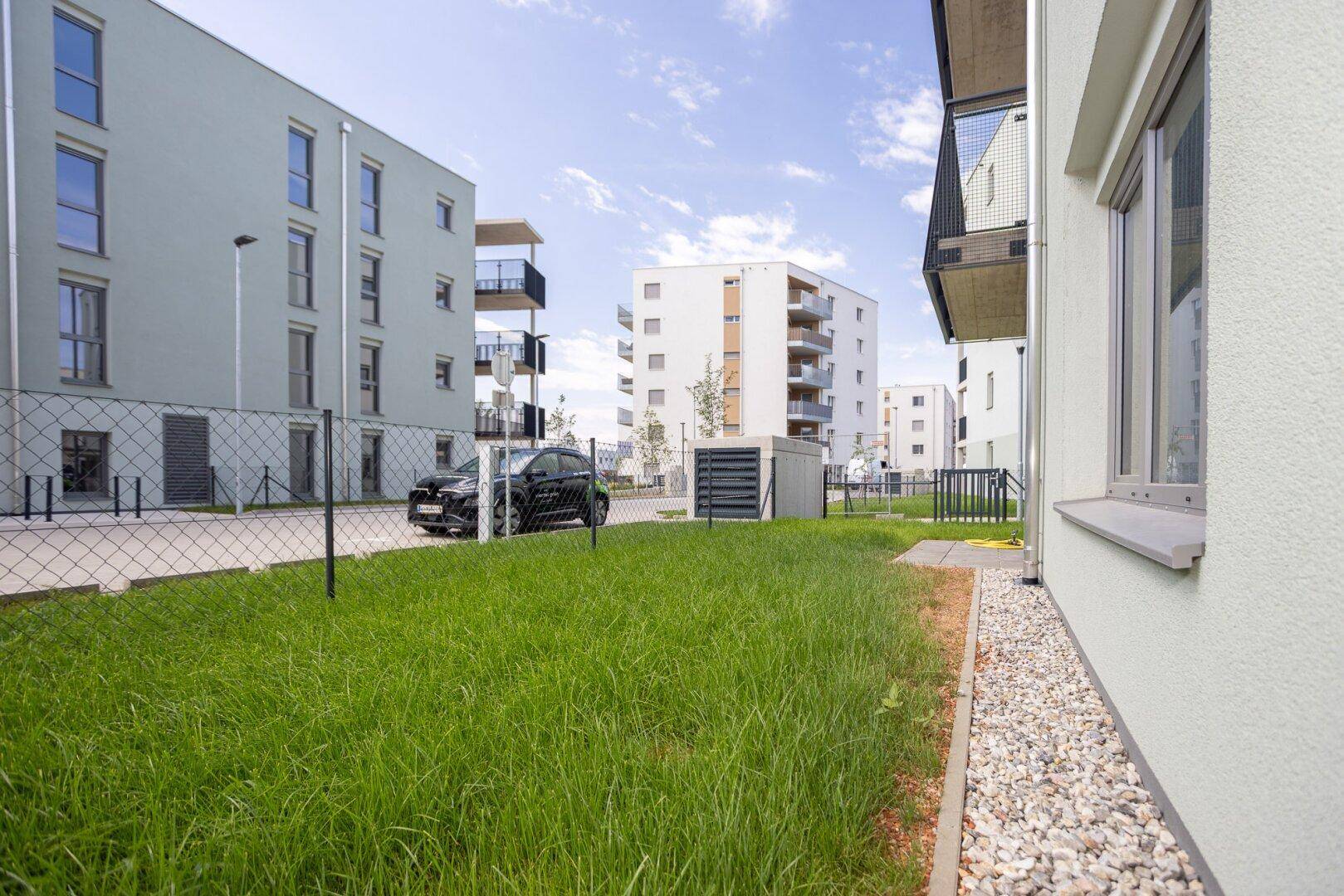 Gepflegte Grünfläche vor modernen Wohngebäuden mit Parkmöglichkeiten und Zaun.