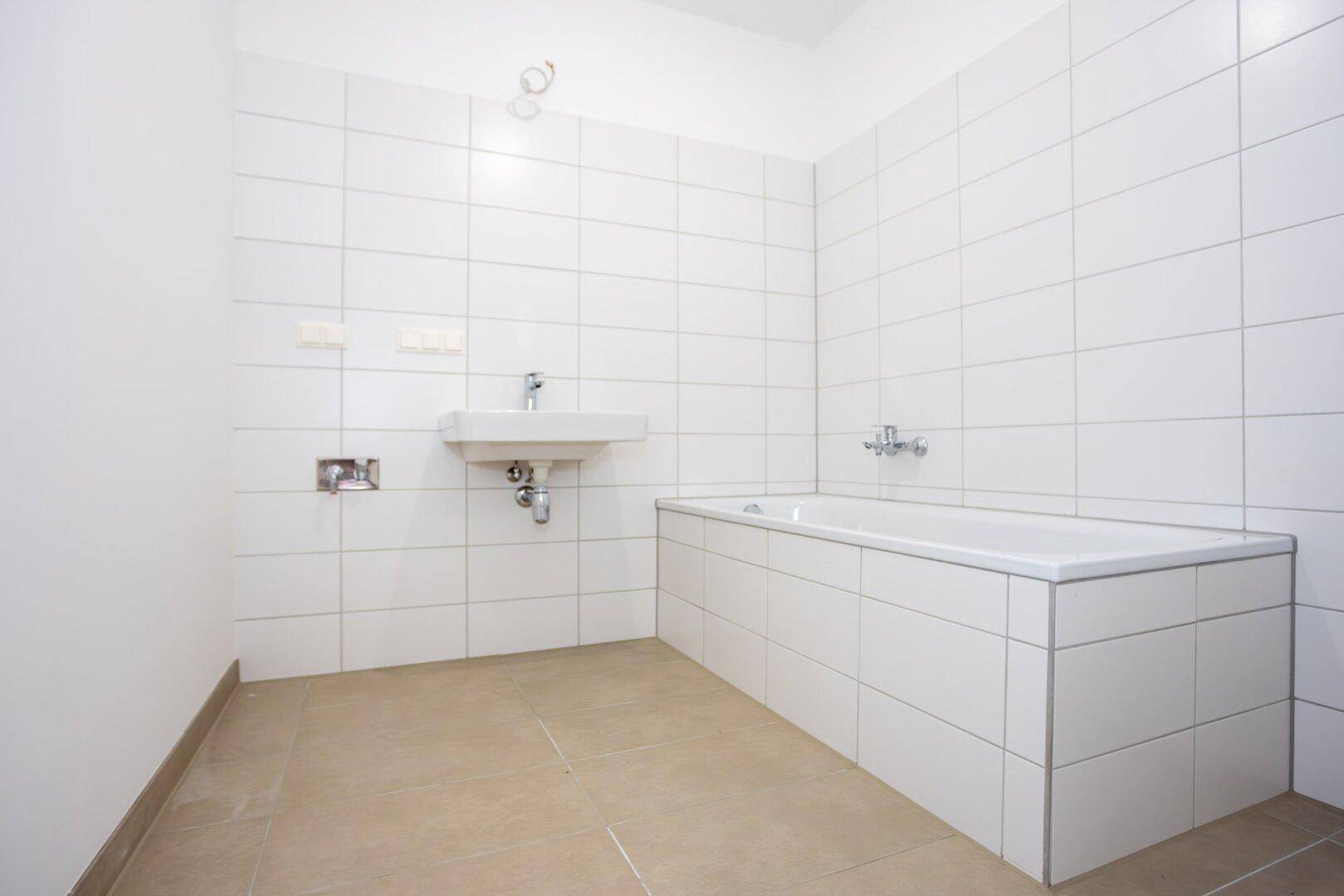 Helles Badezimmer mit weißen Wandfliesen, beigen Bodenfliesen und einer eingebauten Badewanne.