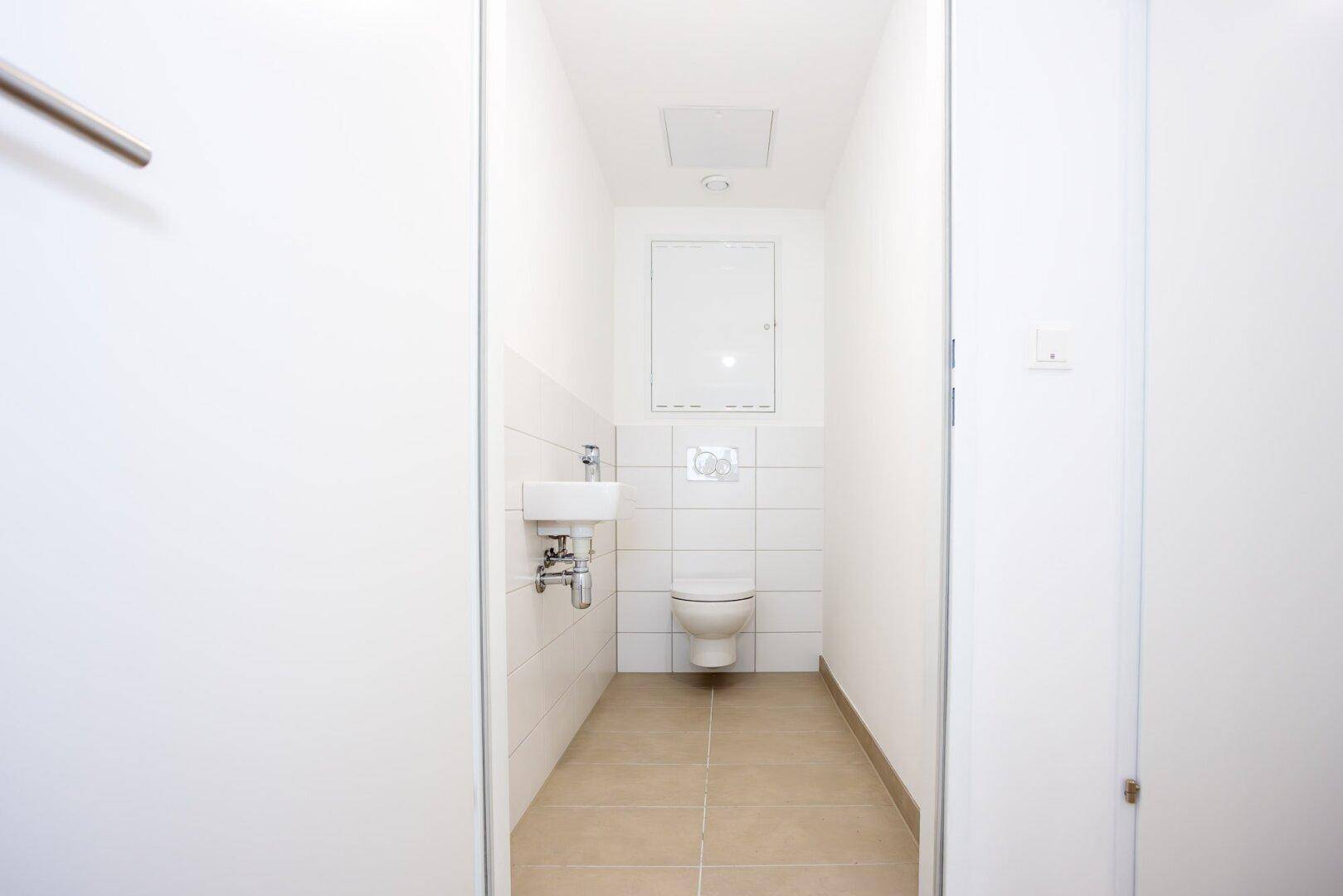 Modernes Gäste-WC mit wandhängender Toilette, kleinem Waschbecken und hellen Fliesen.