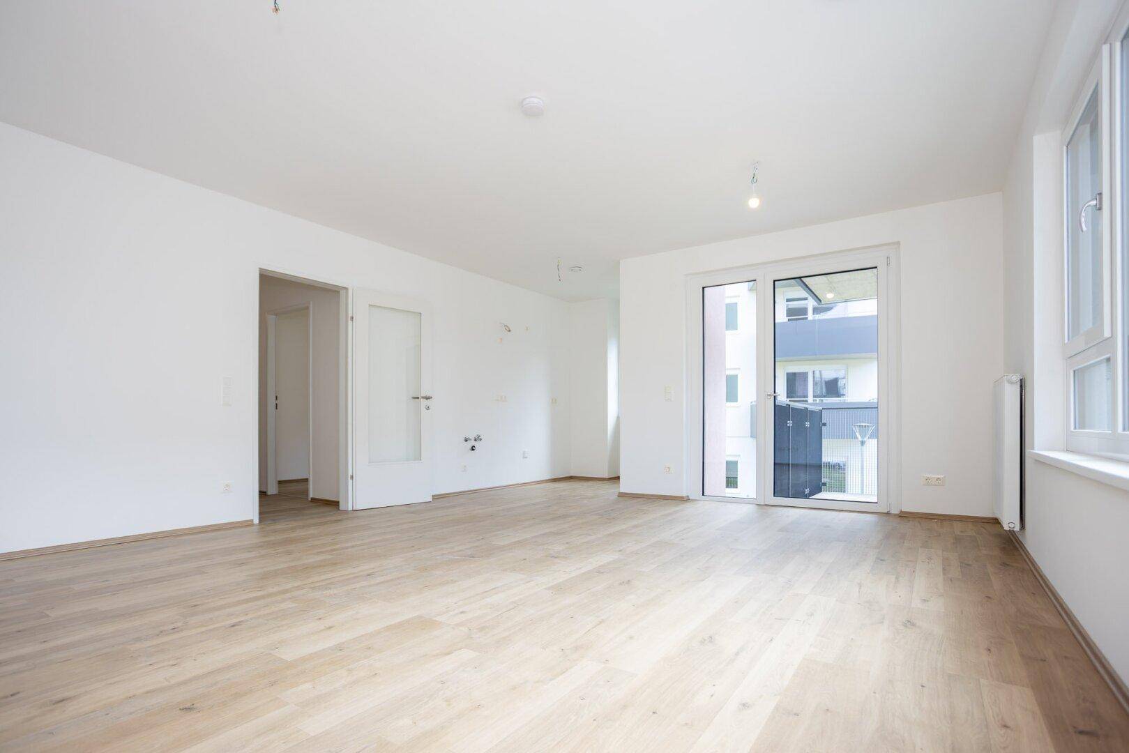 Heller Wohnbereich mit Laminatboden, weißen Wänden und einer Tür, die zu einem weiteren Raum führt.