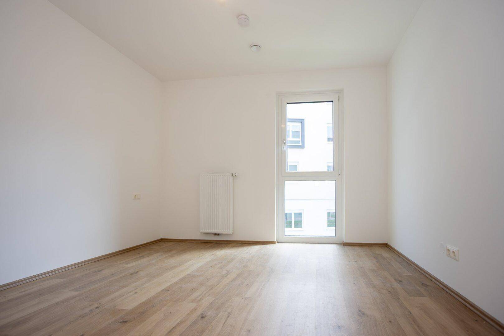 Freundliches Zimmer mit Laminatboden, weißen Wänden und einem Fenster, das viel Tageslicht hereinlässt.