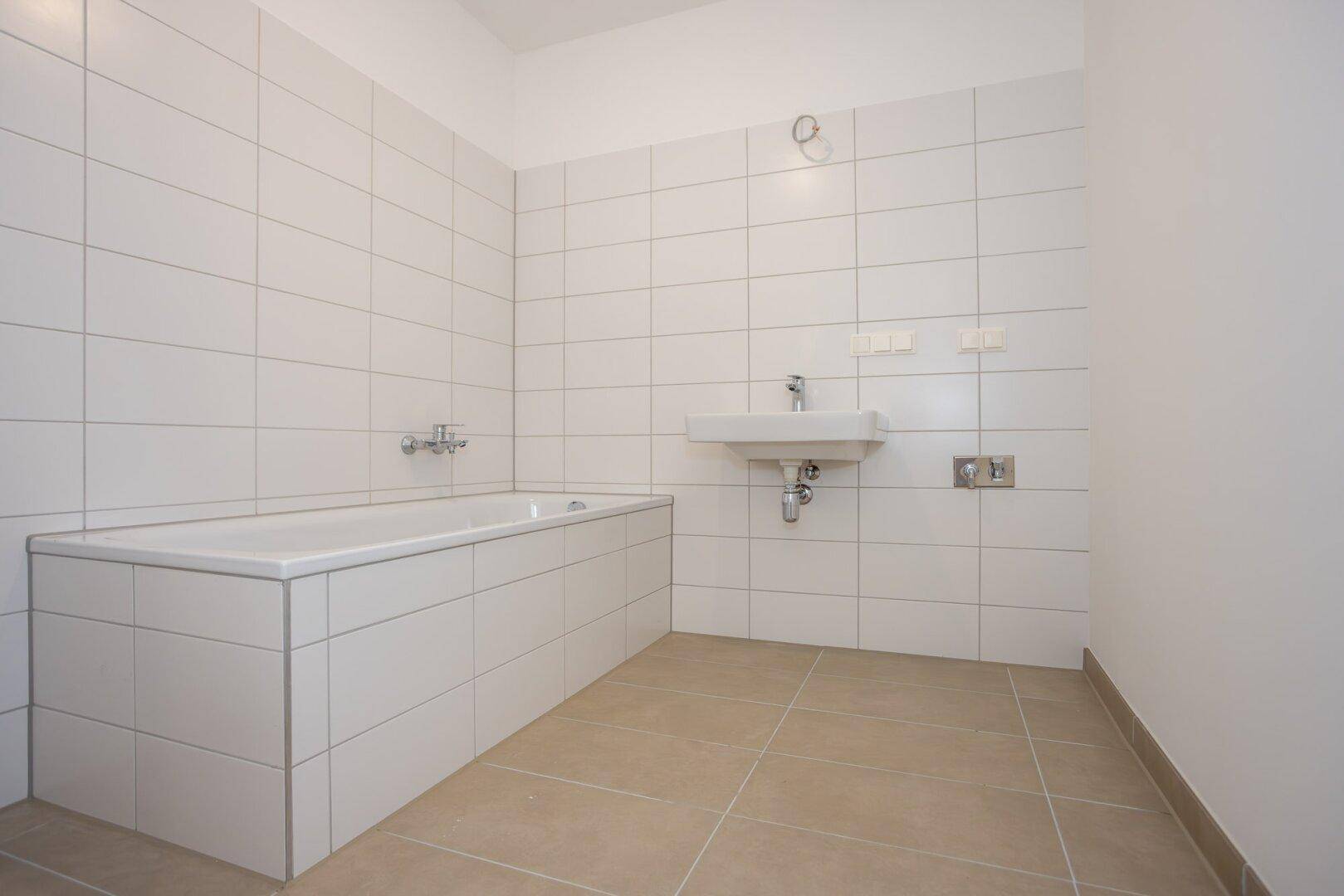 Helles Badezimmer mit eingebauter Badewanne, Waschbecken und weißen Wand- und Bodenfliesen.