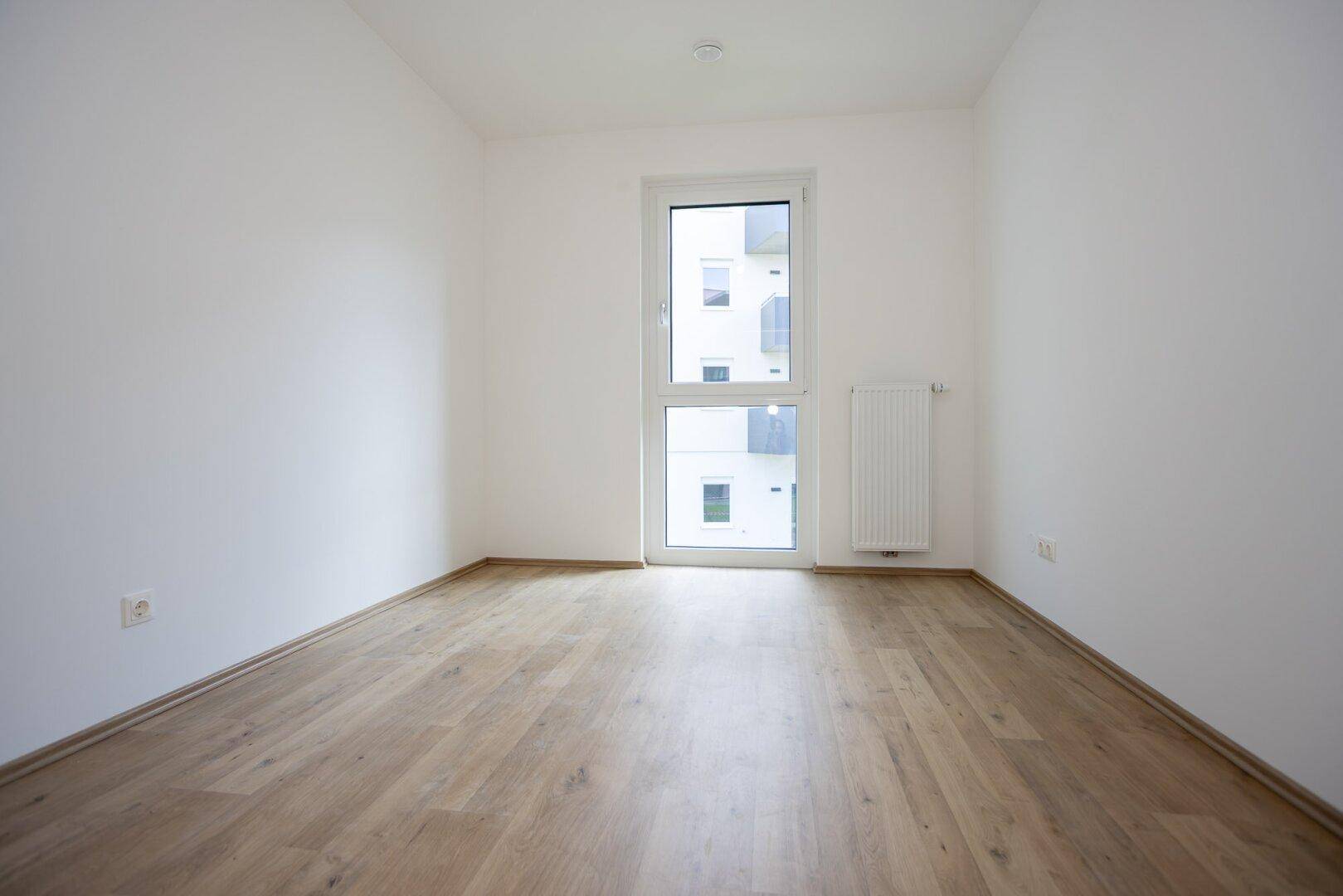 Leeres Schlafzimmer mit hellem Laminatboden, weißen Wänden und einem Fenster für natürliche Belichtung.