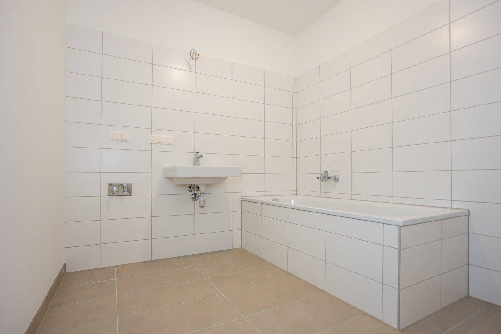 Helles Badezimmer mit Badewanne, Waschbecken und weißen Wand- und Bodenfliesen.